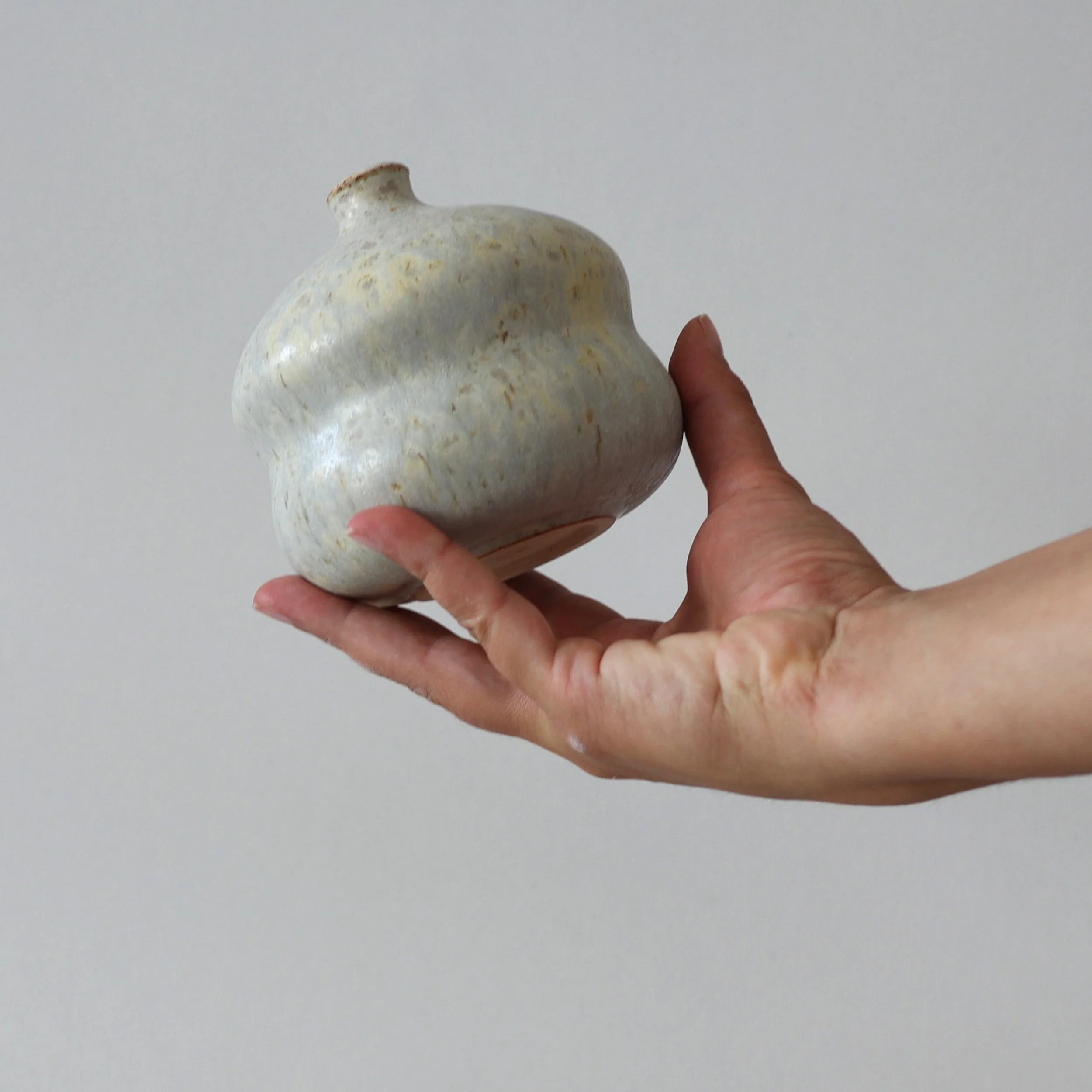 A hand holding a small, rounded, white ceramic pot against a plain gray background.