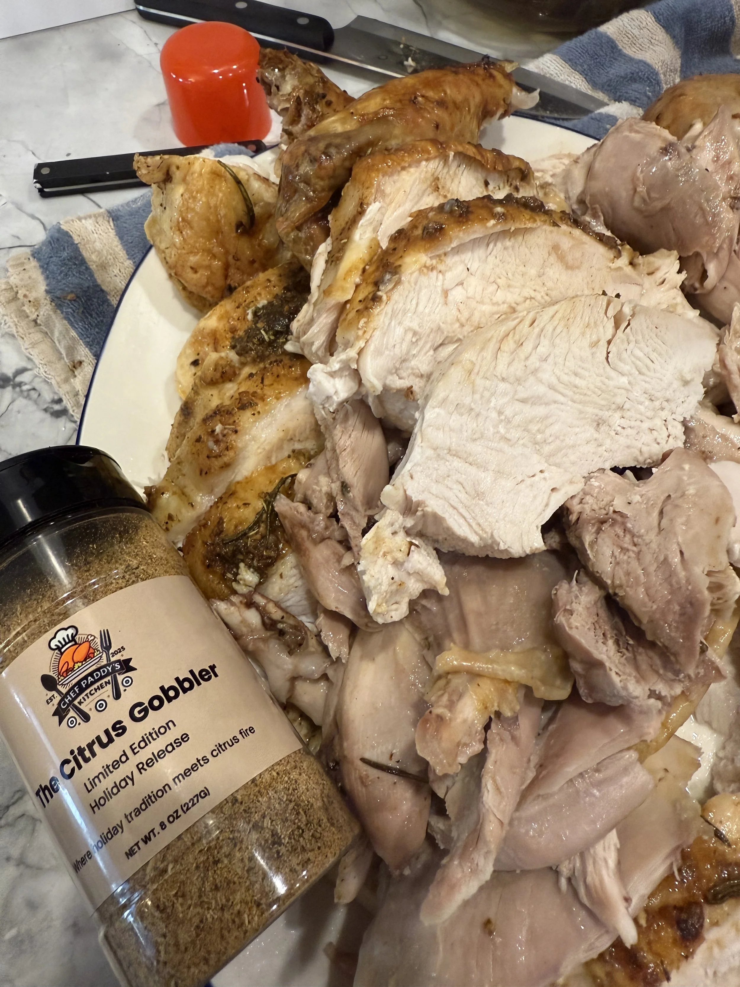 Cooked turkey meat on a cutting board with a jar of Chef Paddy's Citrus Gobbler seasoning, red seasoning shaker, and cutting tools in the background.