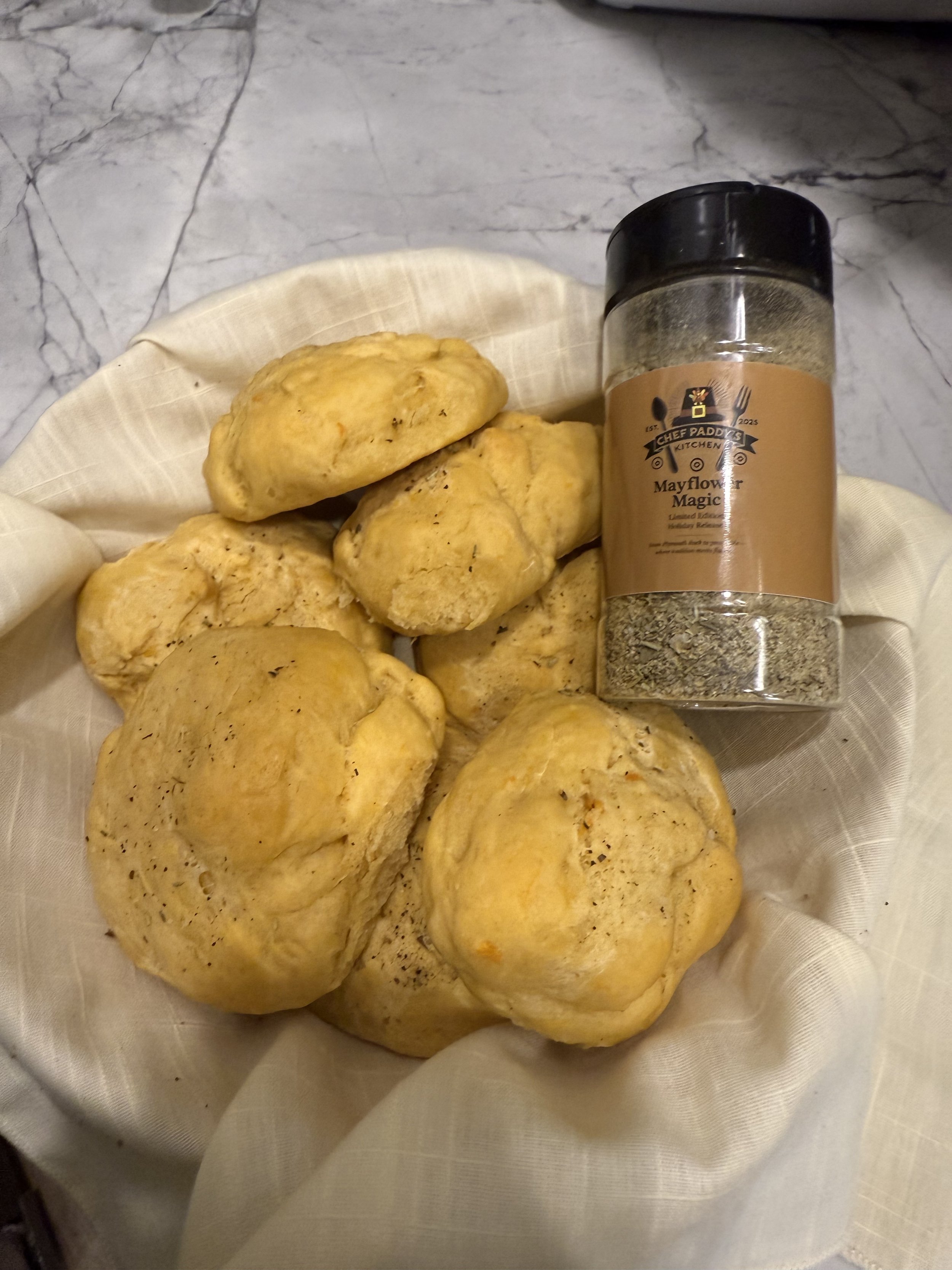 A basket of freshly baked bread rolls with a container of Mayflower Magic seasoning on a marble countertop.