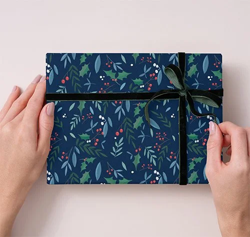 A person ties a black ribbon around a decorated holiday gift box with a blue and green leaf pattern.
