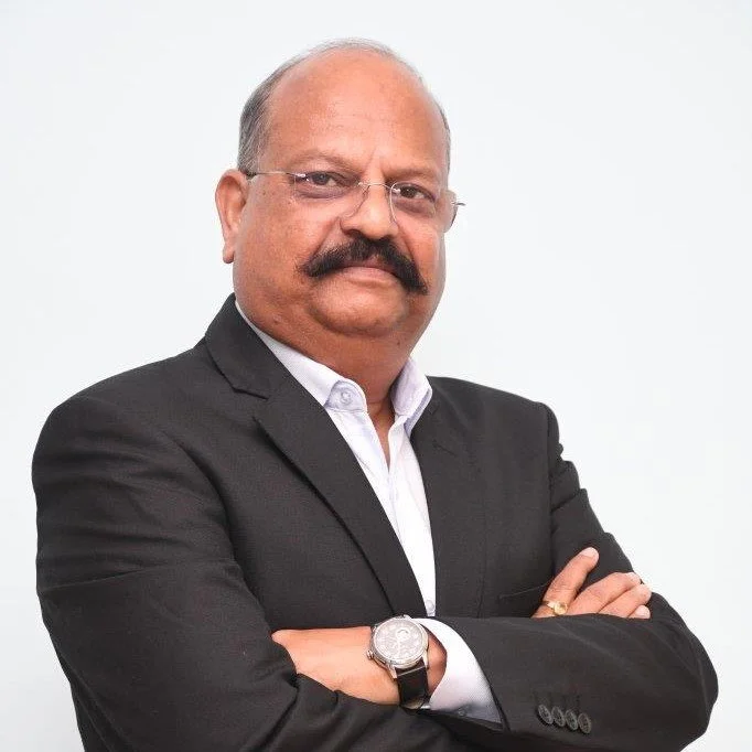 Portrait of a man in a suit with short dark hair and facial hair, wearing a blue shirt and a dark blazer against a light background.