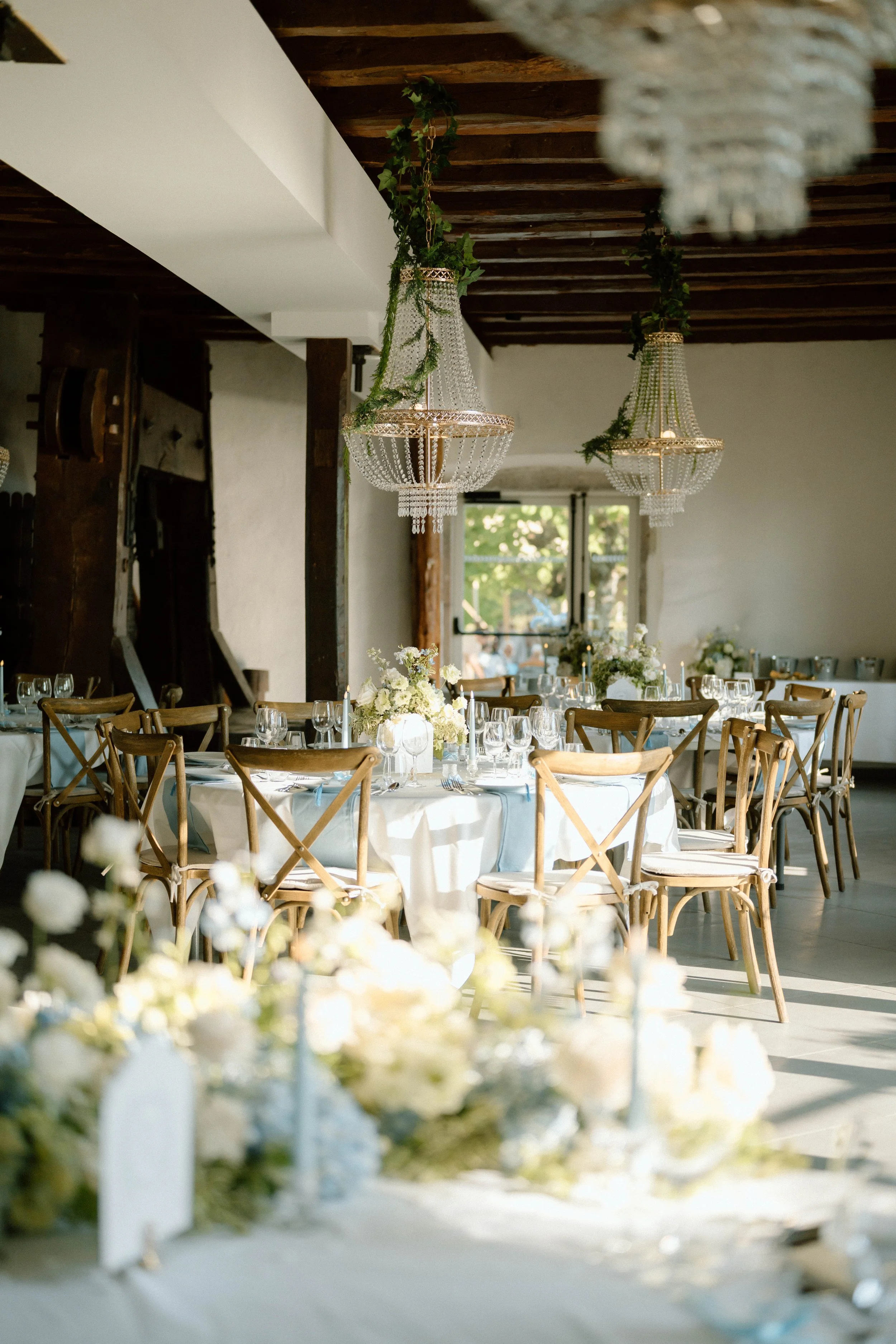 Salle de réception décorée avec des chandeliers en cristal suspendus, des tables rondes avec nappes blanches, des fleurs blanches en centre de table et une grande fenêtre laissant entrer la lumière naturelle.