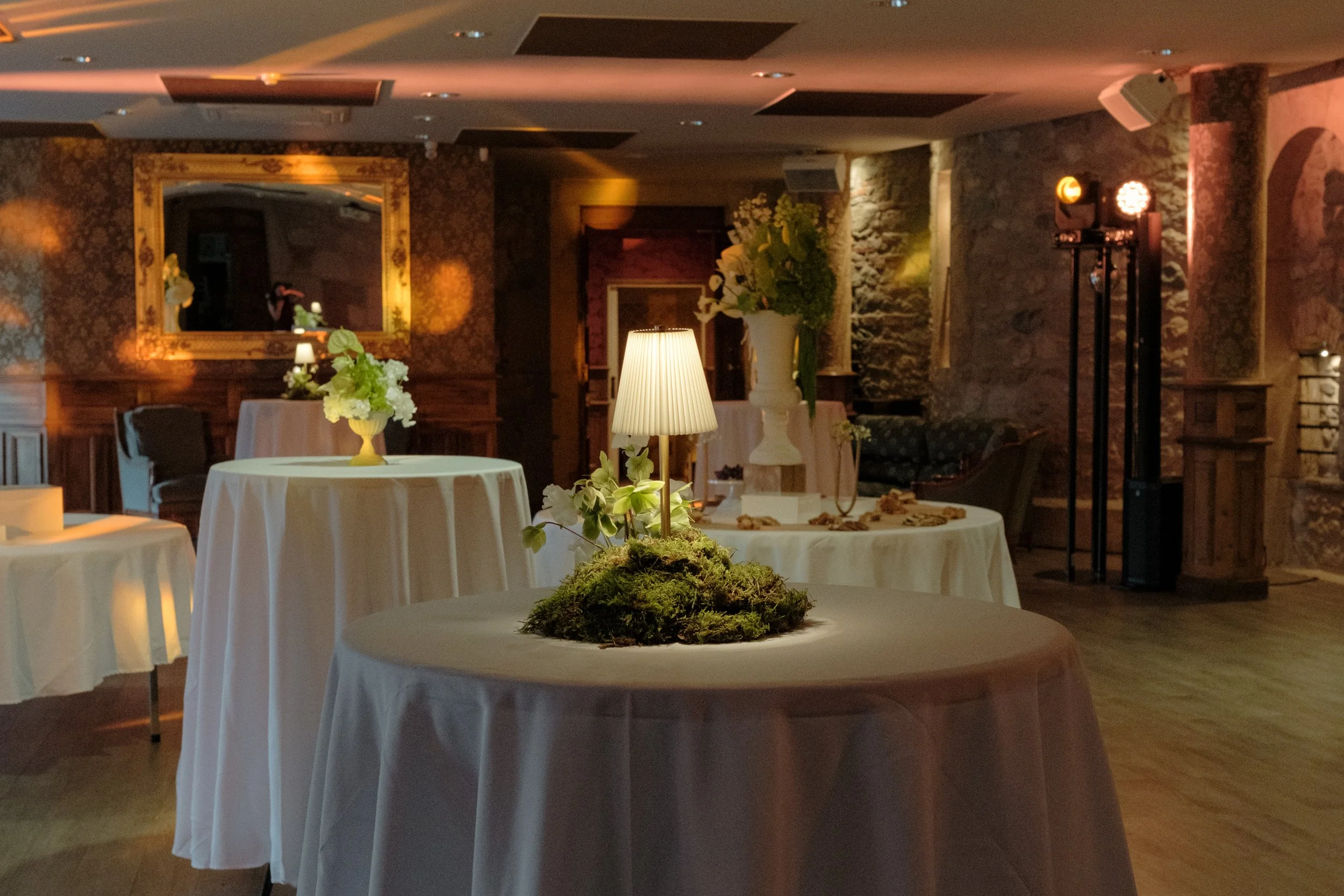 Salle de réception élégante avec des tables rondes drapées de nappe blanche, décorées de centres de fleurs et d'une petite lampe. Mur en pierre, décor avec miroir orné et éclairage doux.
