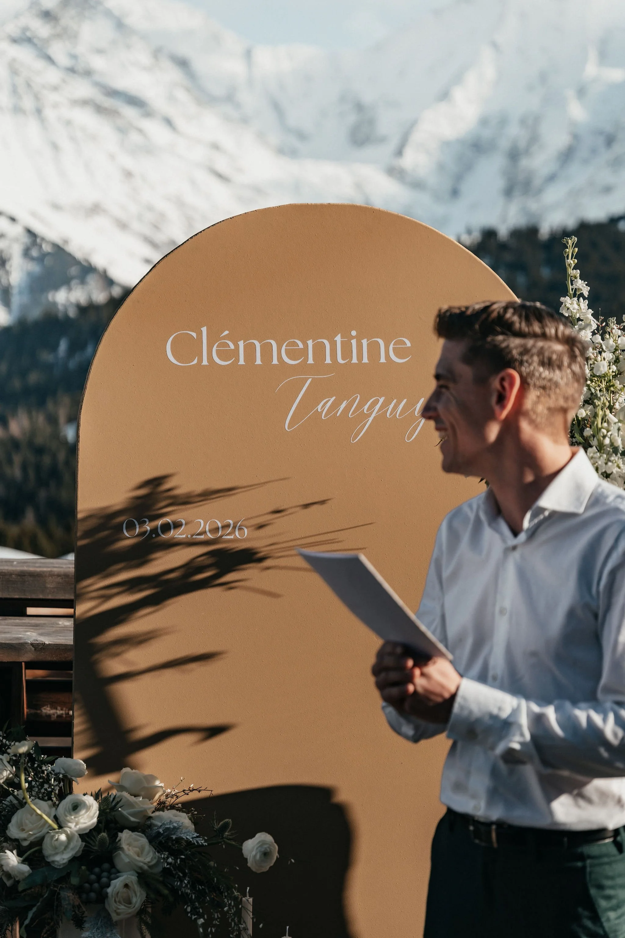 Un homme souriant éclaire un discours lors d'un mariage en plein air, avec montagnes enneigées en arrière-plan et un panneau beige avec le nom Clémentine Tanguy et la date 03.02.2026.