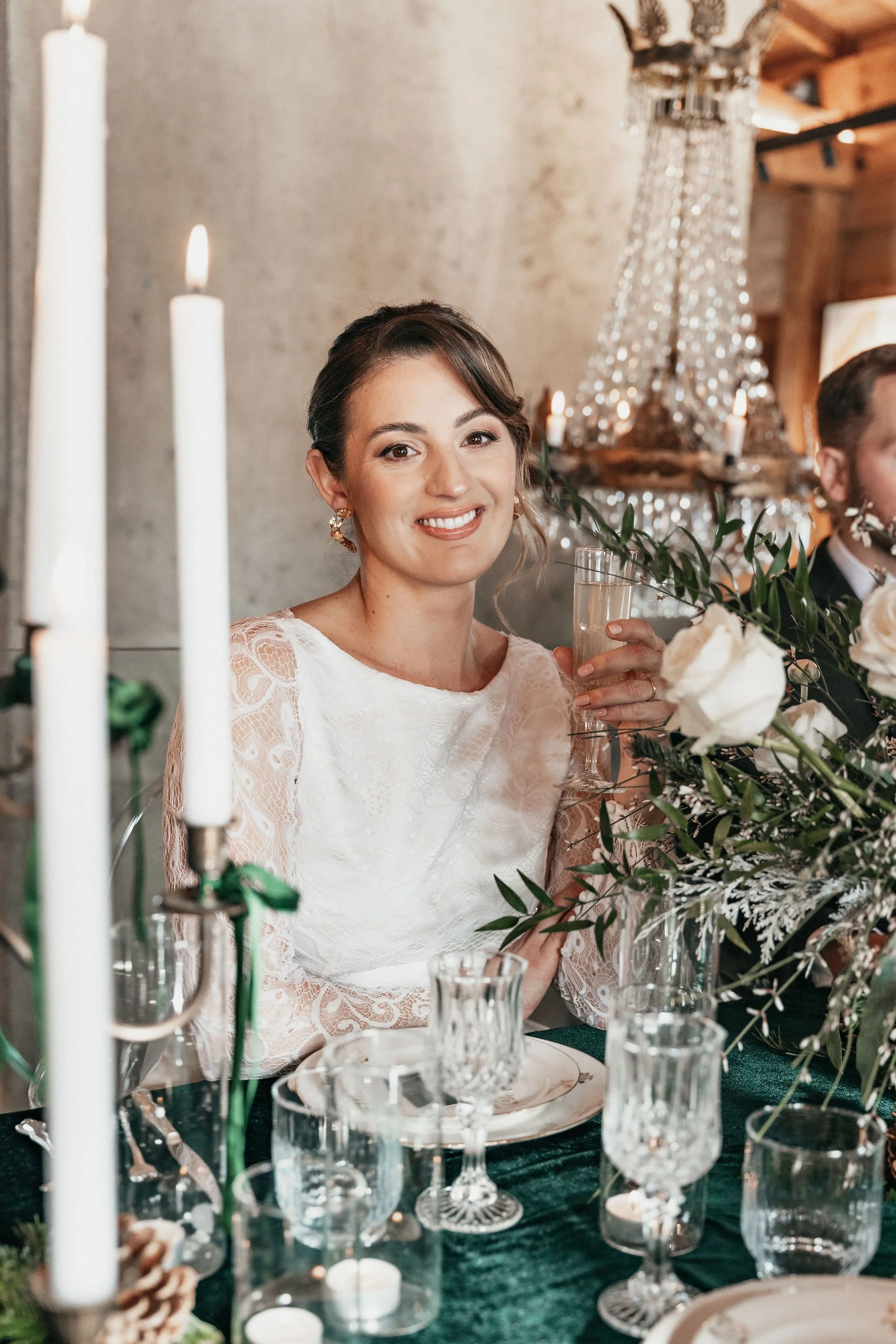 Une femme souriante à un mariage, portant une robe en dentelle blanche, tenant un verre de champagne. La table est décorée avec des bougies et des fleurs.