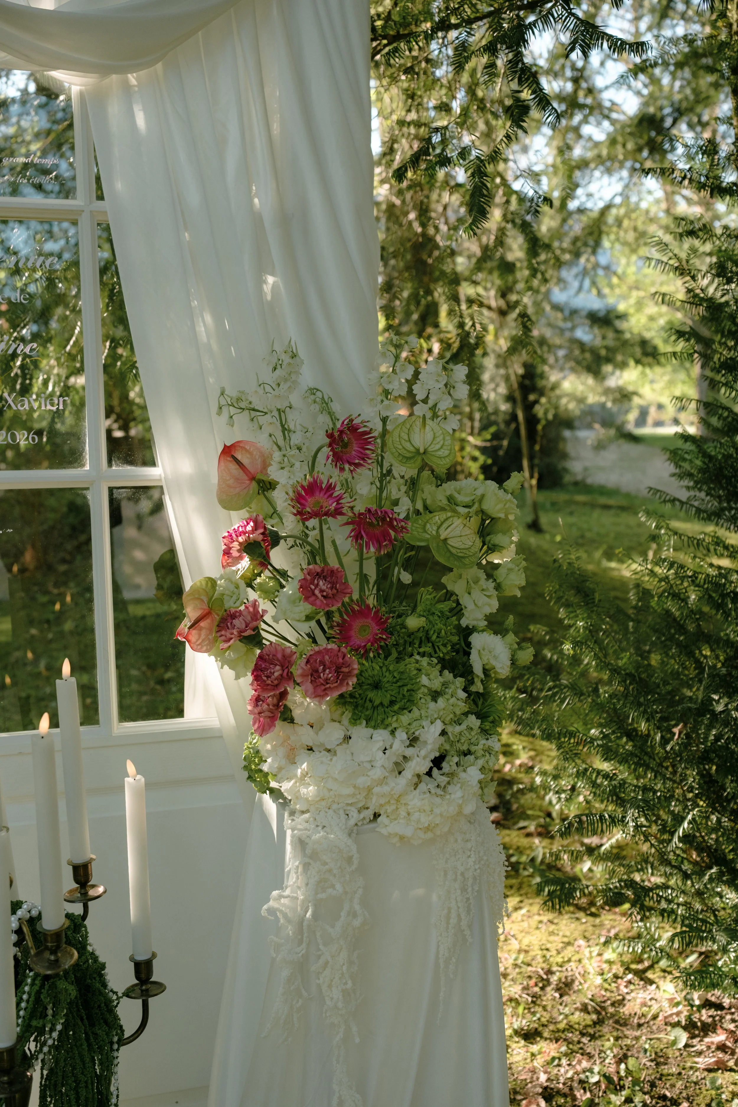 Une décoration florale pour un mariage avec un paravent blanc, des fleurs blanches et roses, de bougies et un environnement extérieur verdoyant.