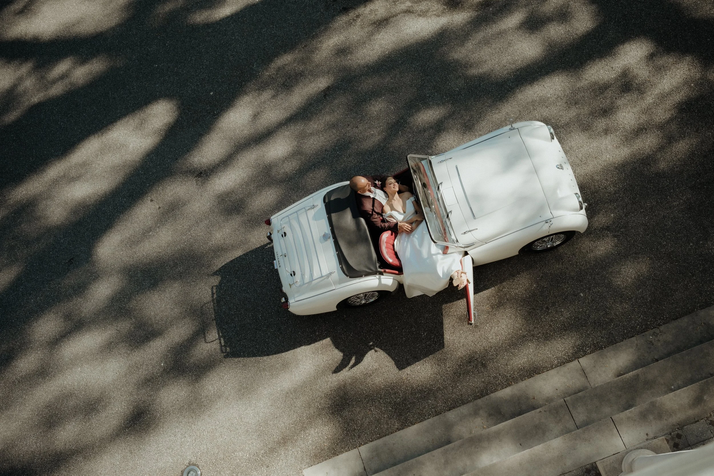 Une voiture blanche décapotable avec un couple à l'intérieur, vu du dessus, sur un sol en pierre avec des ombres de feuilles.