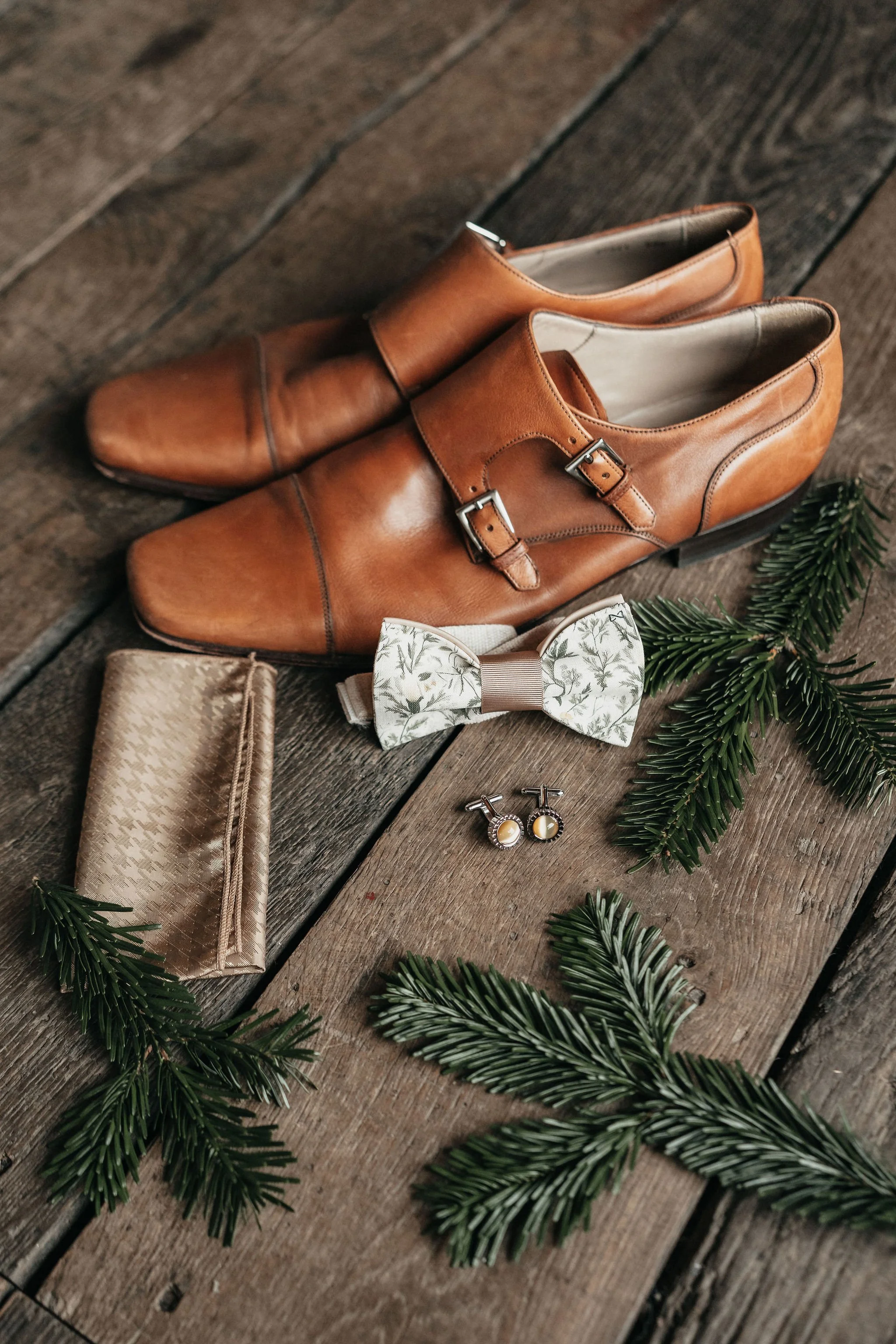 Chaussures en cuir marron, nœud papillon avec motif floral, une pochette beige, boutons de manchette et branches de sapin sur un sol en bois.