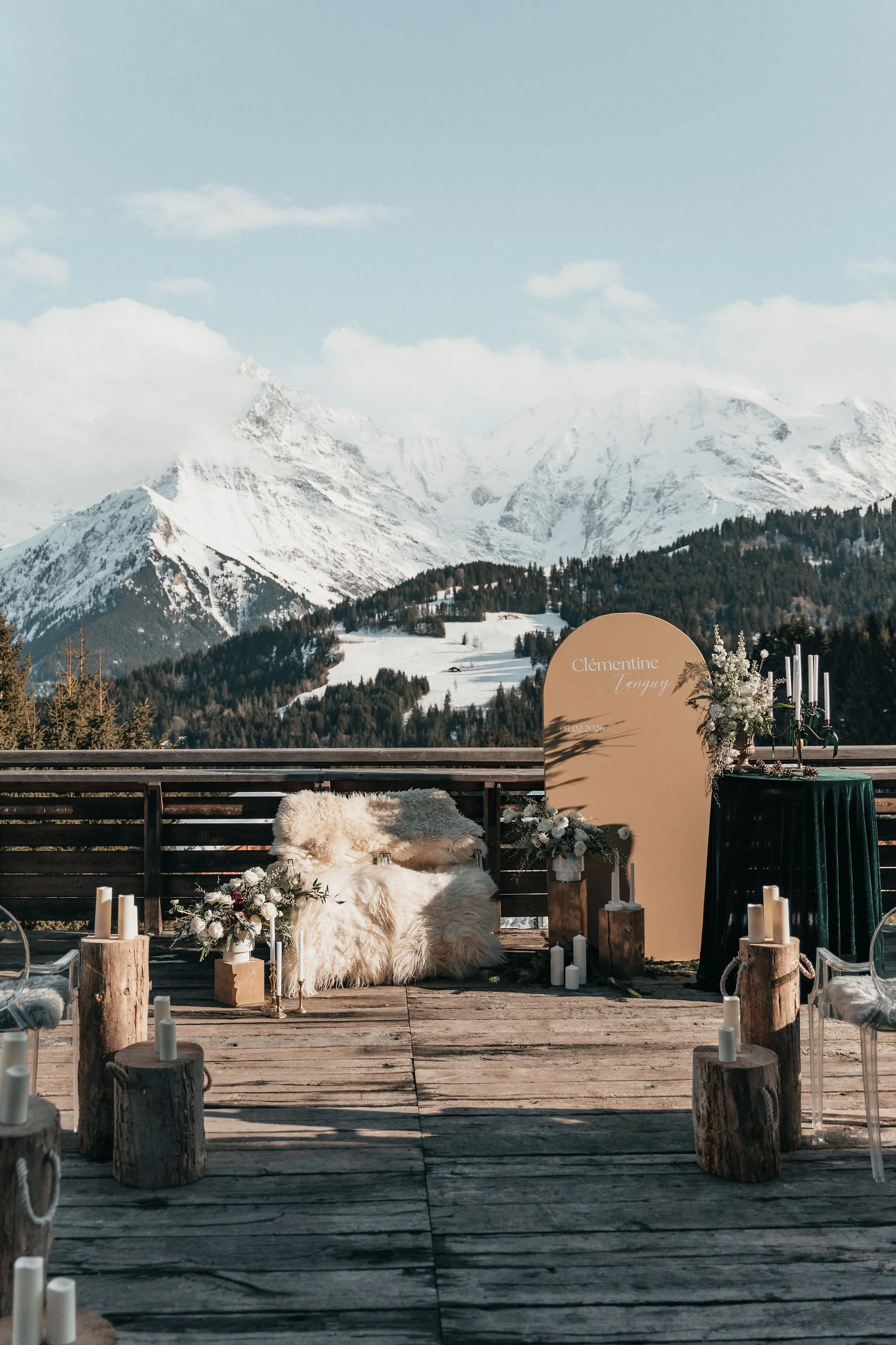 Décoration de mariage en montagne avec un décor en bois, fleurs blanches, bougies, et un panneau avec le nom Clémentine Tanguy, en extérieur avec des montagnes enneigées en arrière-plan.