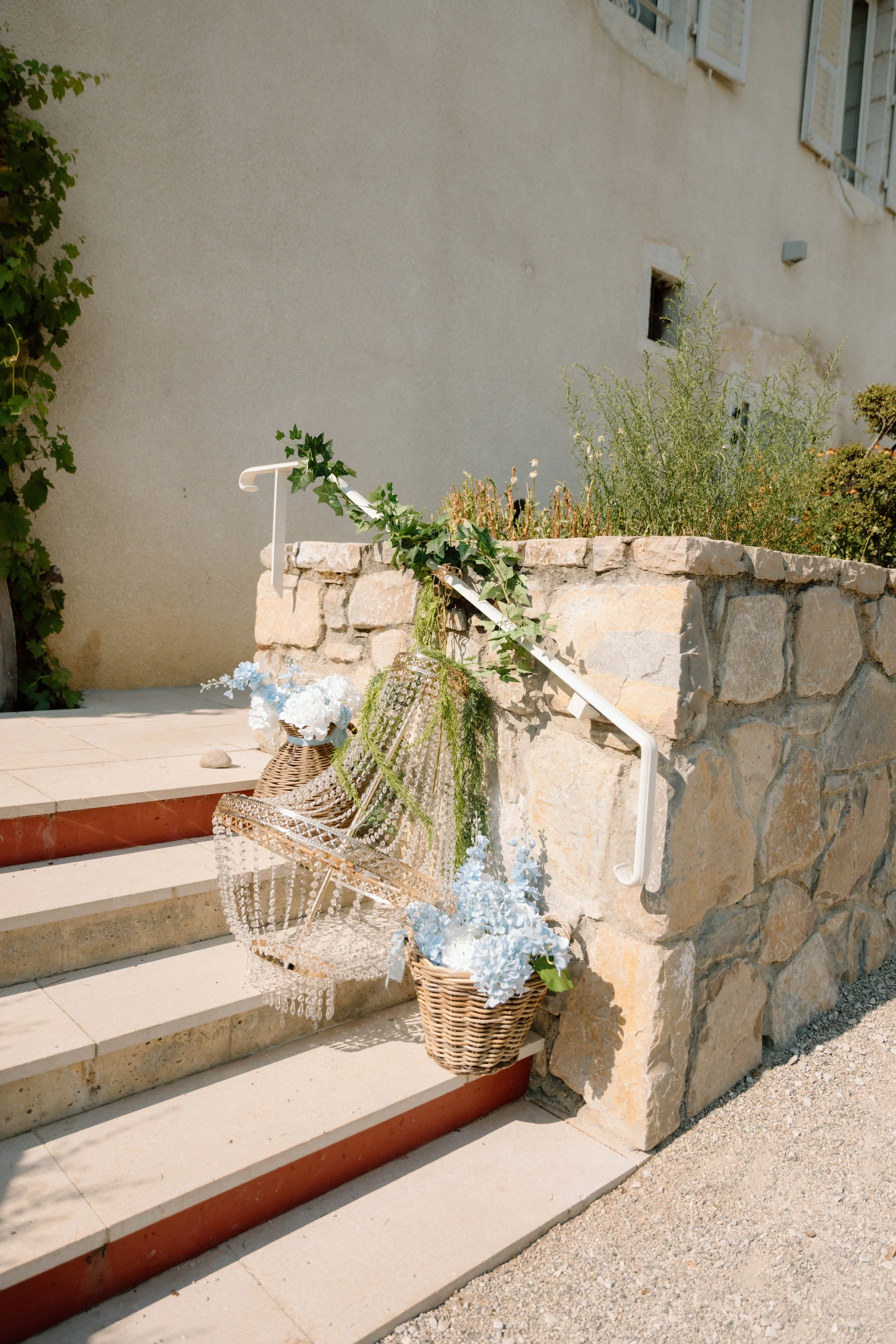 Escalier en pierre menant à une maison avec des fenêtres à volets bleus, décoré avec des paniers de fleurs et une statue décorative. Parterres de plantes à l'arrière.