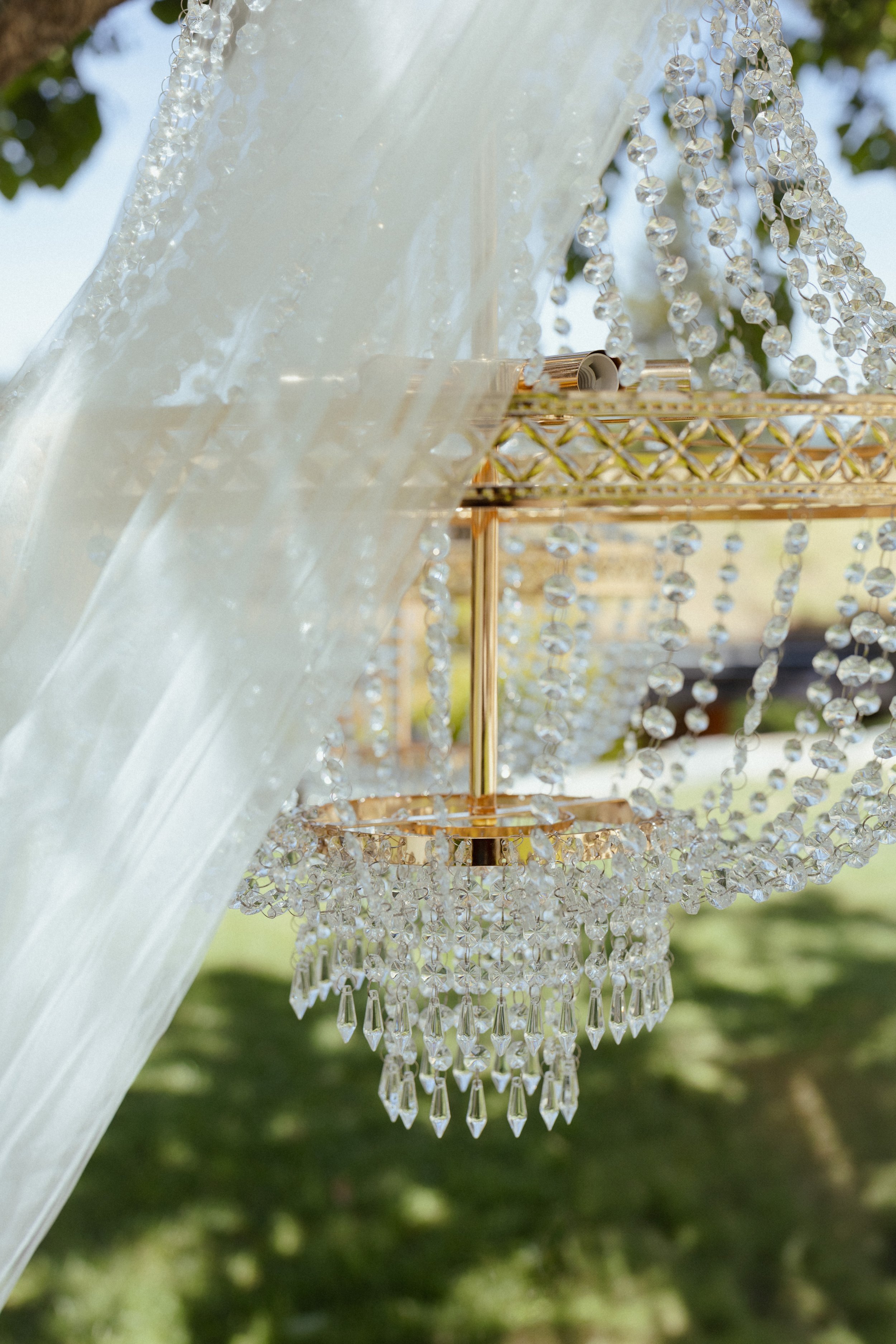 Lustre en cristal suspendu avec des chaînes de cristaux scintillants, côté extérieur avec un fond flou de végétation verte.
