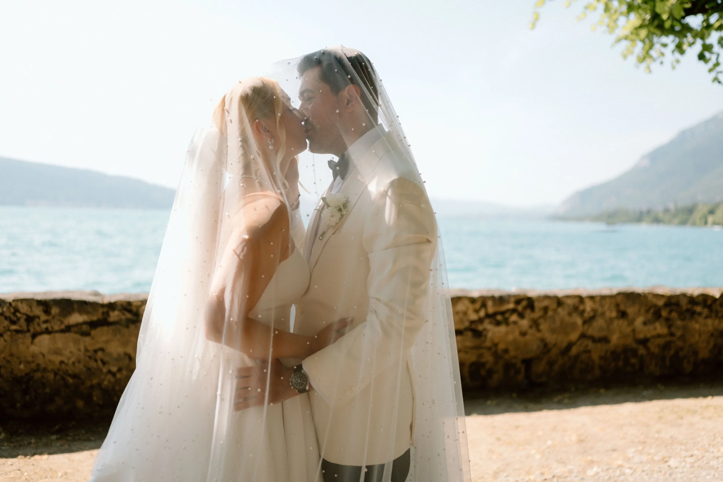 Un couple en habits de mariage se embrassant sous un voile de mariée, avec un lac et des montagnes en arrière-plan.