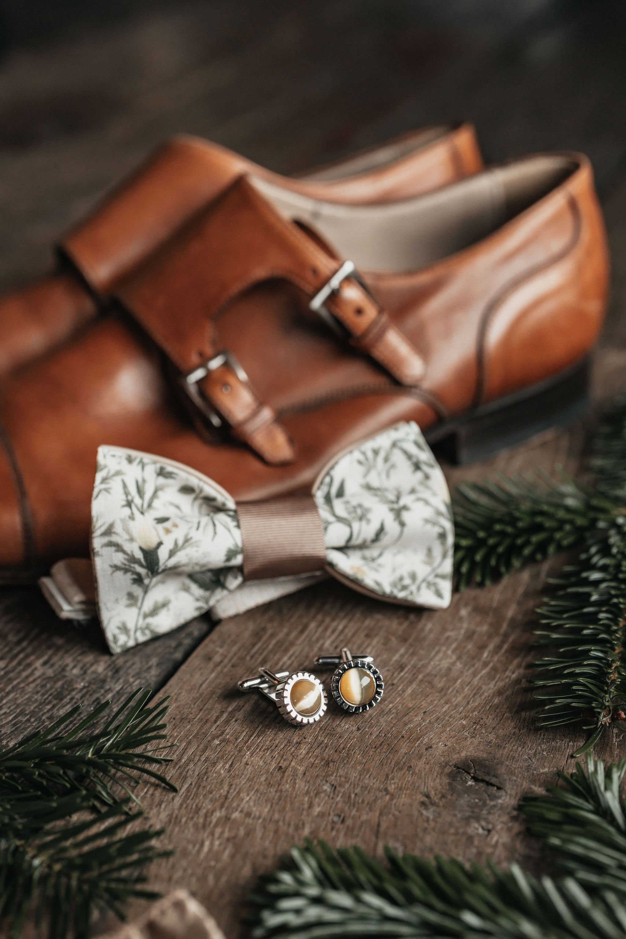 Une paire de chaussures en cuir marron, une mouchette papillon en tissu avec un motif floral, une paire de boutons de manchette avec un design en forme d'œuf, des branches d'épinette et un sac en cuir en arrière-plan.