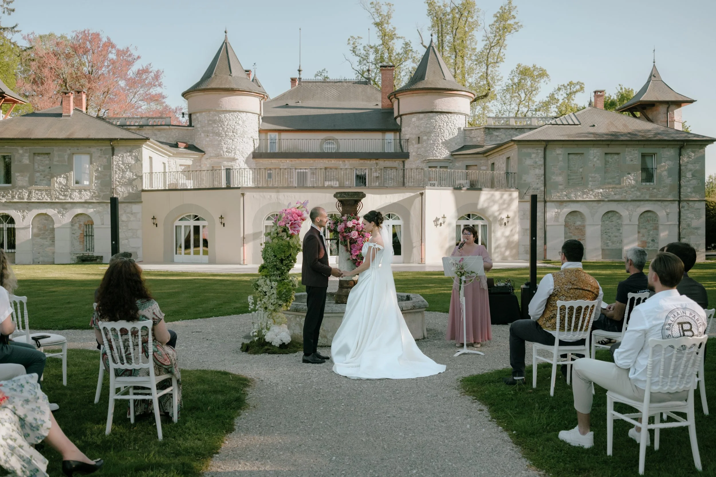 Un mariage en plein air devant un bâtiment historique avec deux tours en pierre et un toit en pente. La mariée et le marié échangent leurs vœux, entourés de leurs invités assis en plein air.