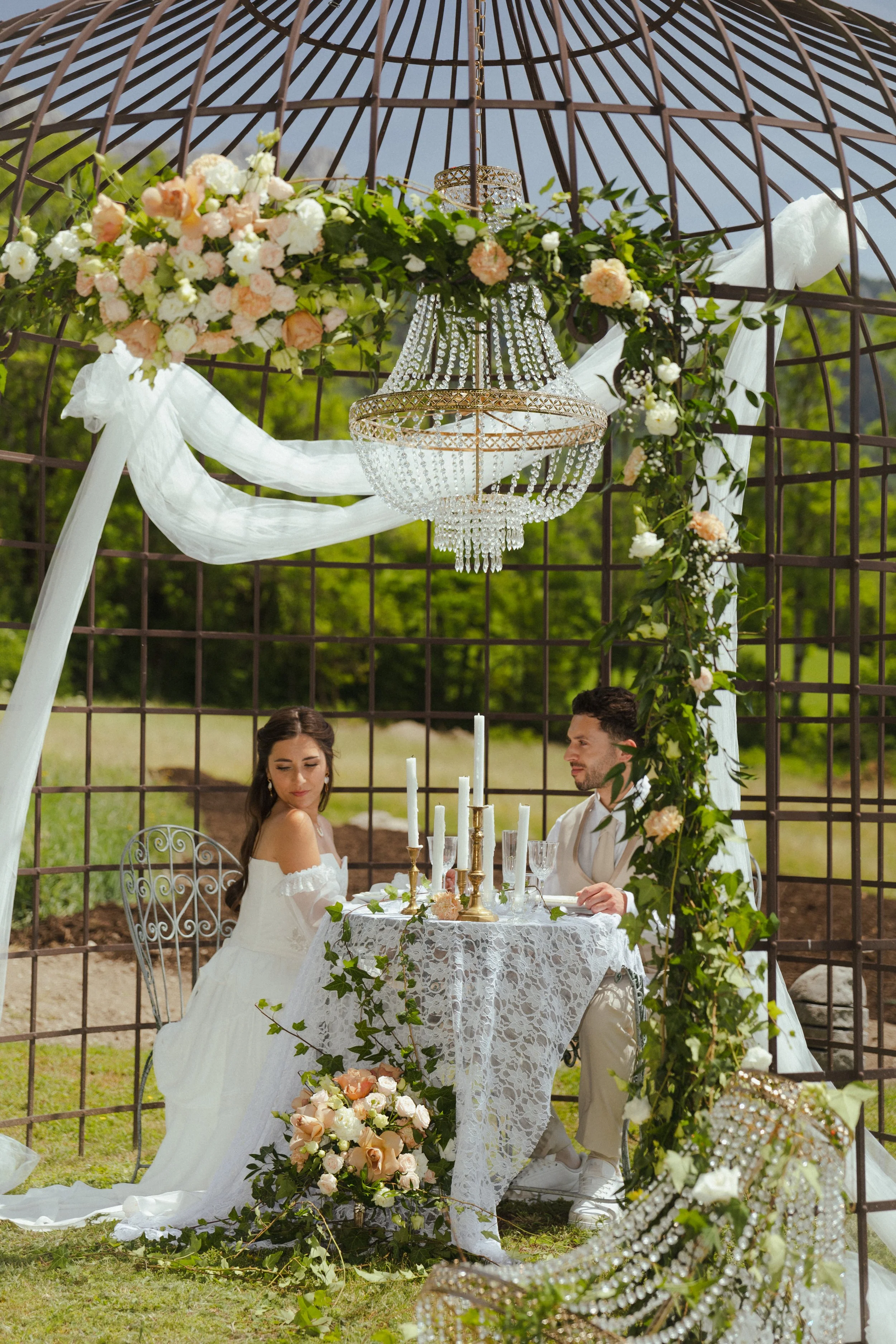 Un couple assis à une table de mariage en extérieur, sous une arche décorée de fleurs et drapé blanc, avec un chandelier en cristal au centre, lors d'une cérémonie nuptiale.