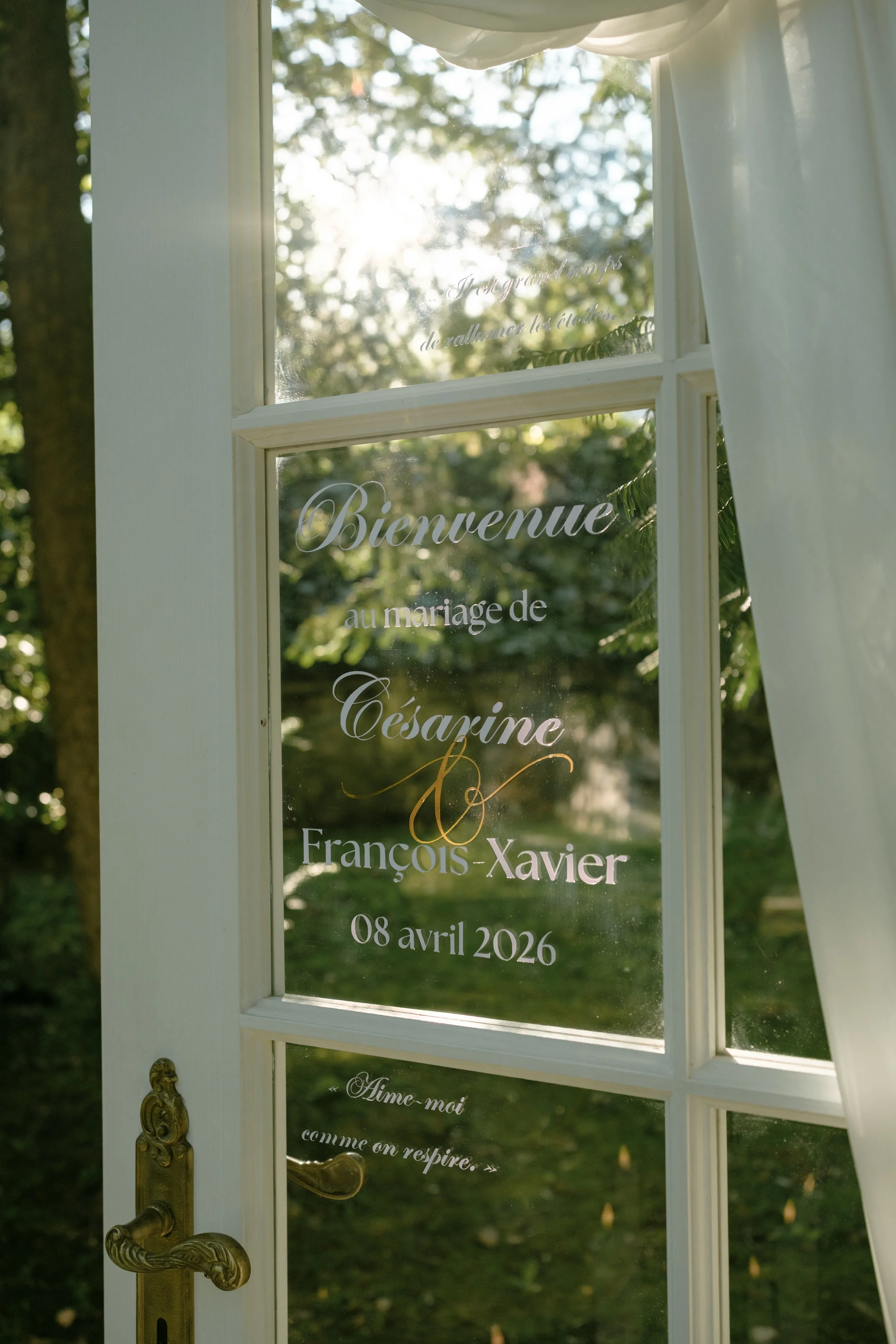 Une porte en verre avec des inscriptions en français, notamment "Bienvenue", une date, et des noms, derrière une fenêtre avec une vue sur un jardin avec des arbres et la lumière du soleil.