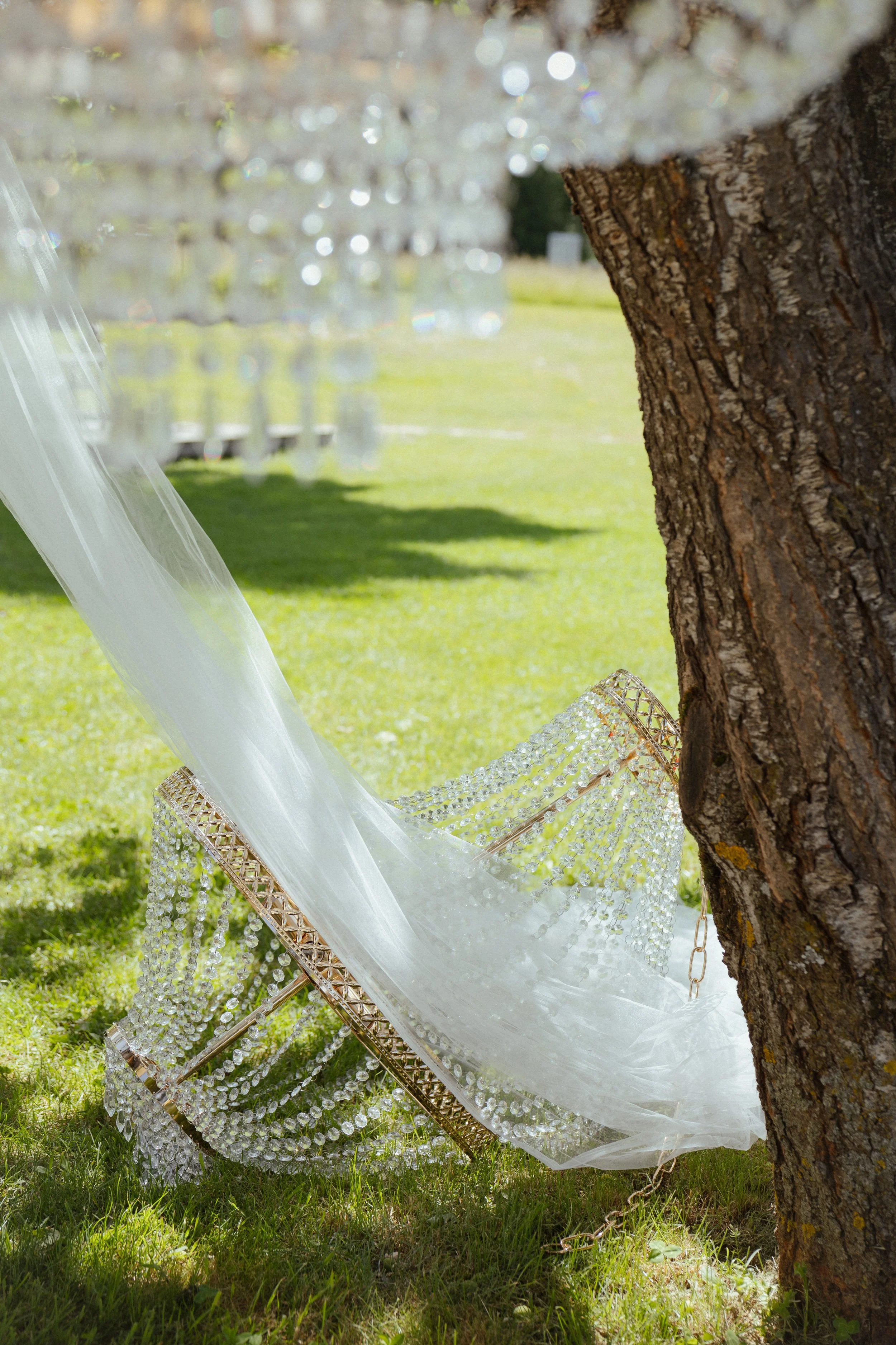 Une chaise pliante en métal doré est décorée de perles et de tissu blanc, positionnée sous un arbre avec un tronc rugueux, dans un espace extérieur avec de l'herbe verte et un fond flou de lumières lumineuses