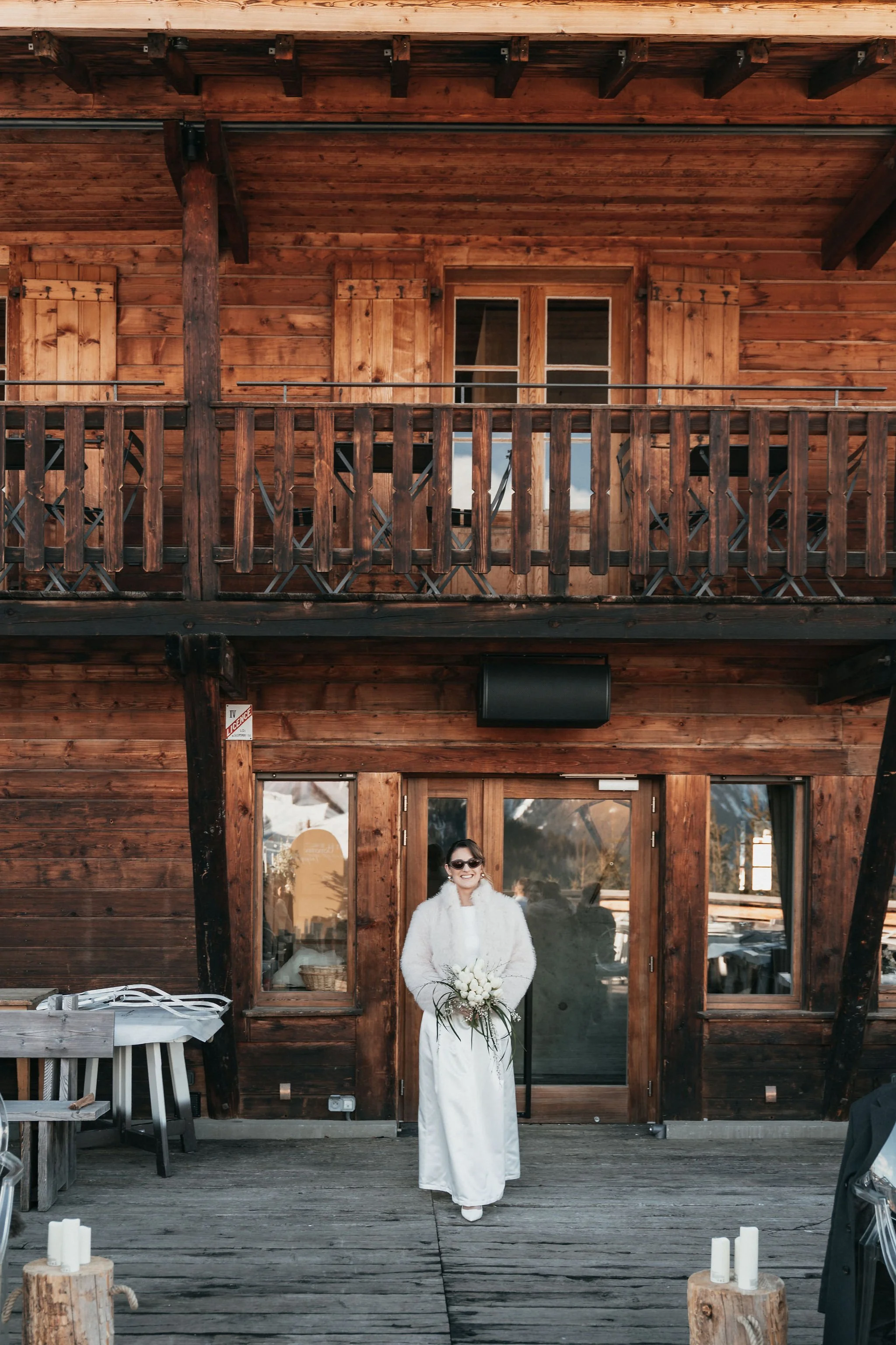 Une femme en robe blanche et manteau blanc, avec des lunettes de soleil, tenant un bouquet de fleurs, se tenant devant une façade en bois d'un bâtiment avec balcon. Photo prise en extérieur.