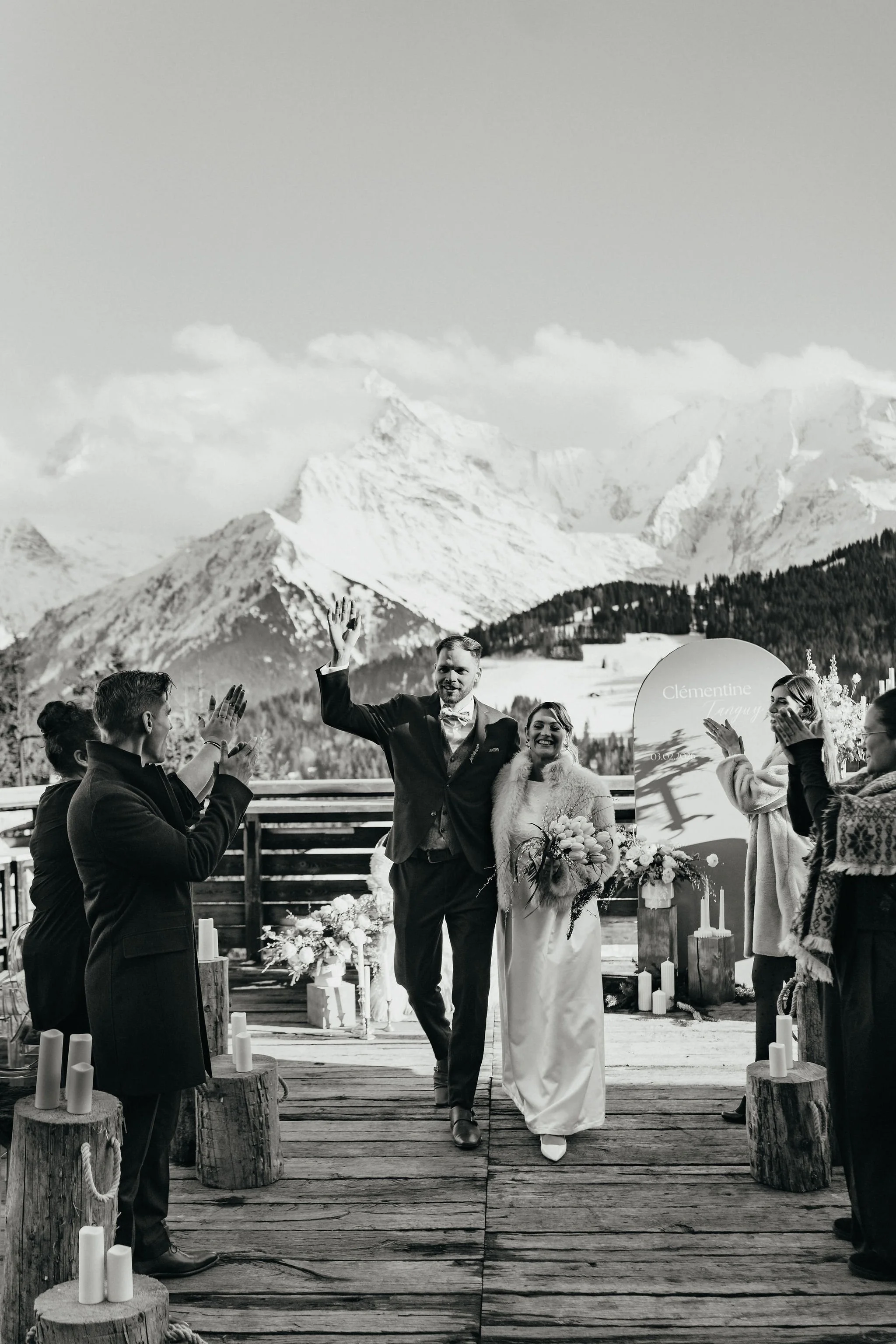 Un couple de mariés marche main dans la main lors de leur mariage dans un lieu en montagne, entouré d'amis qui les applaudissent.