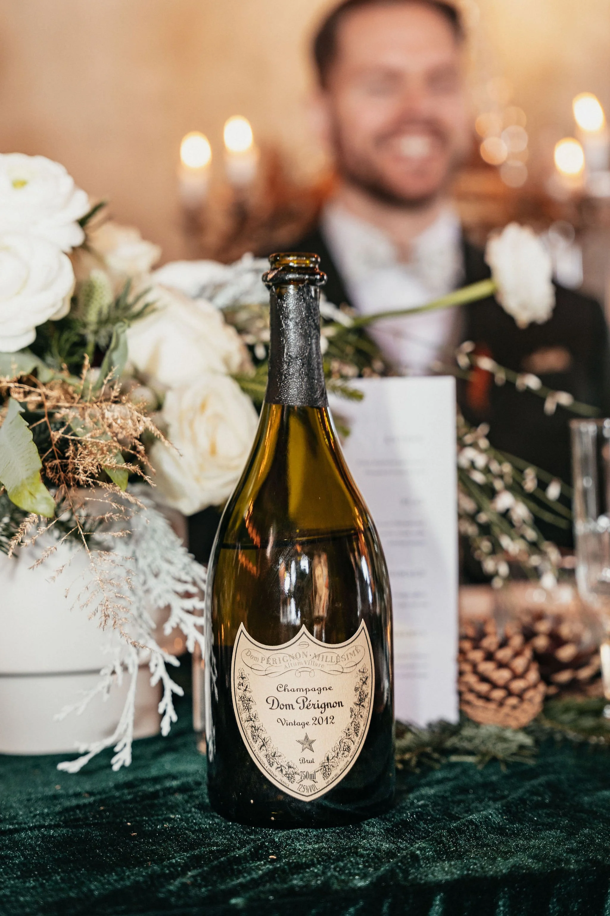 Bouteille de champagne Dom Pérignon Vintage 2012, décor de fête avec fleurs blanches et bougies en arrière-plan.