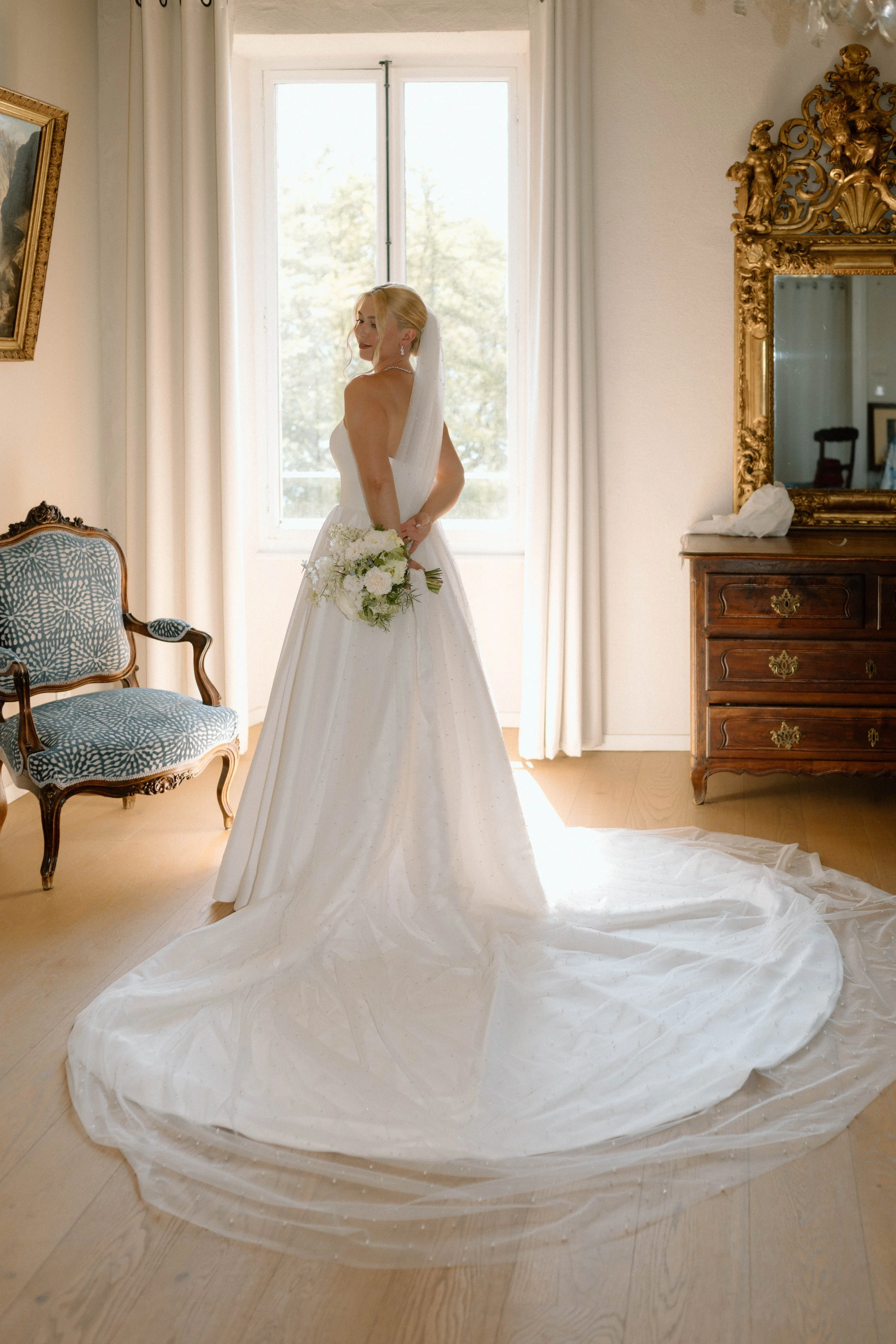 Une mariée en robe de mariée blanche tenant un bouquet de fleurs blanches, se tenant devant une fenêtre dans une pièce élégante avec un grand miroir doré et un fauteuil ancien.