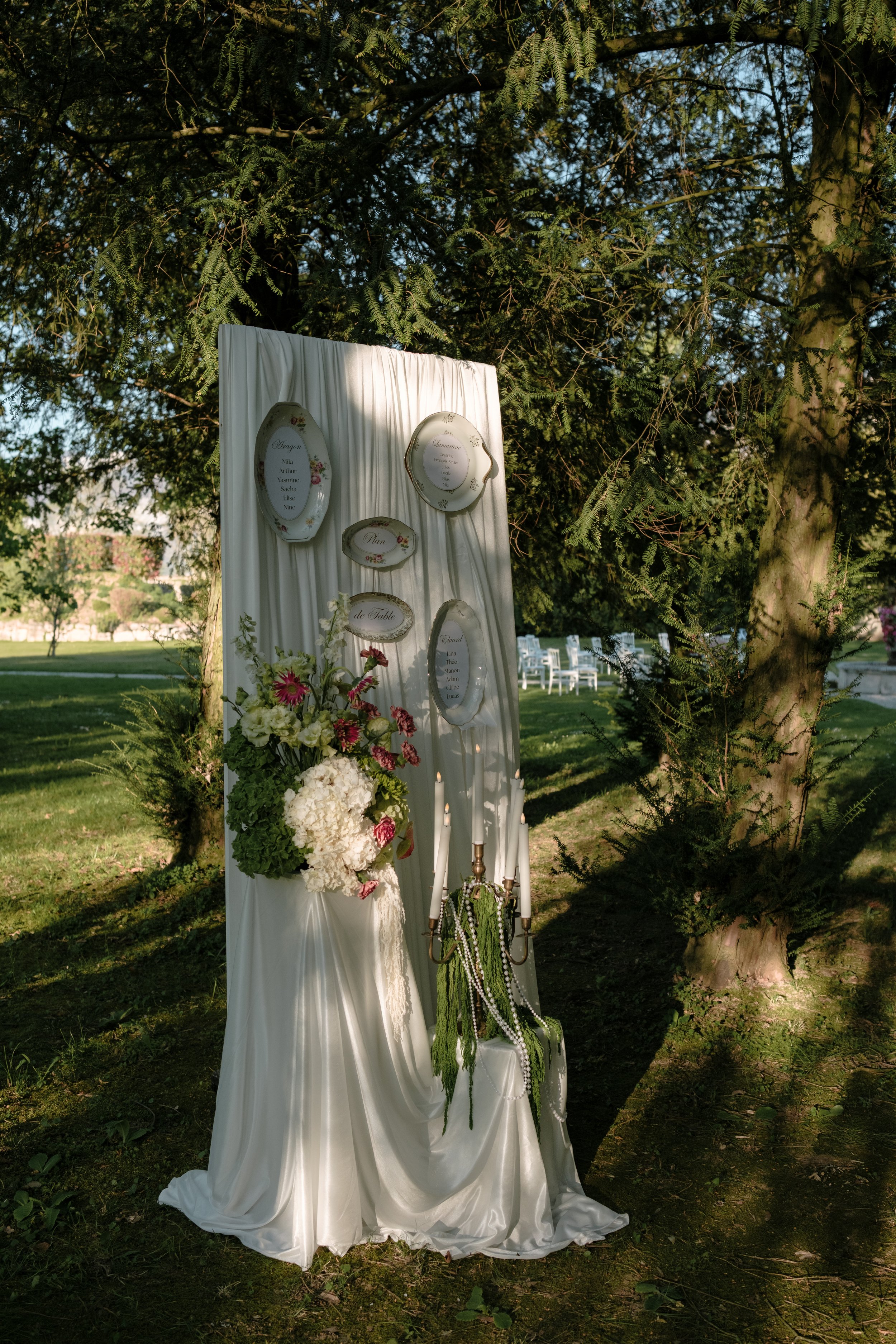 Décoration de mariage en plein air avec un panneau blanc et des assiettes suspendues, fleurs blanches et roses, bougies et chandelier à l'extérieur à l'ombre des arbres.
