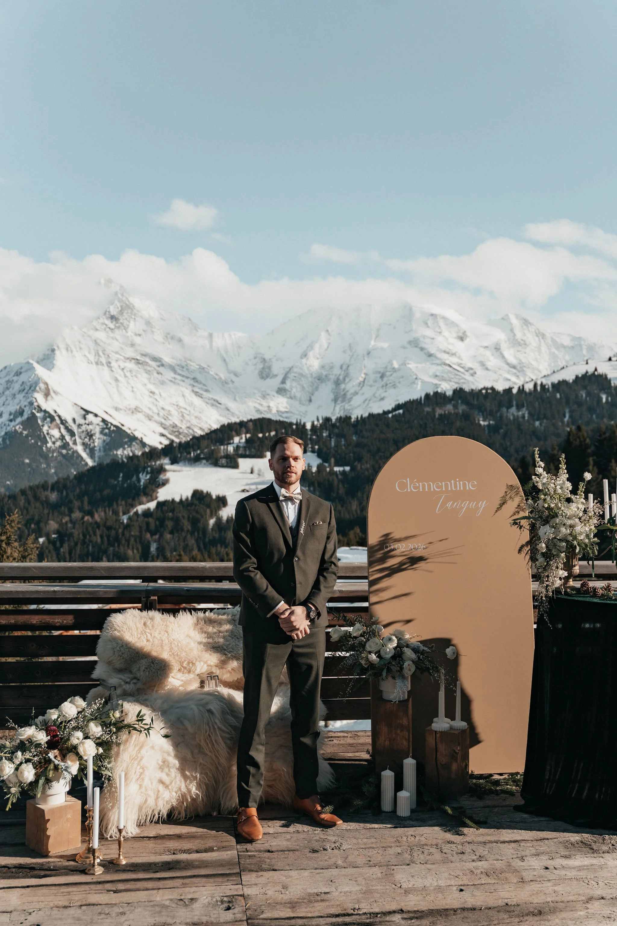 Un homme habillé en costume dans un décor de mariage en extérieur avec montagnes enneigées en arrière-plan, décorations florales et bougies autour d'une grande arche portant le nom Clémentine Tanguy.