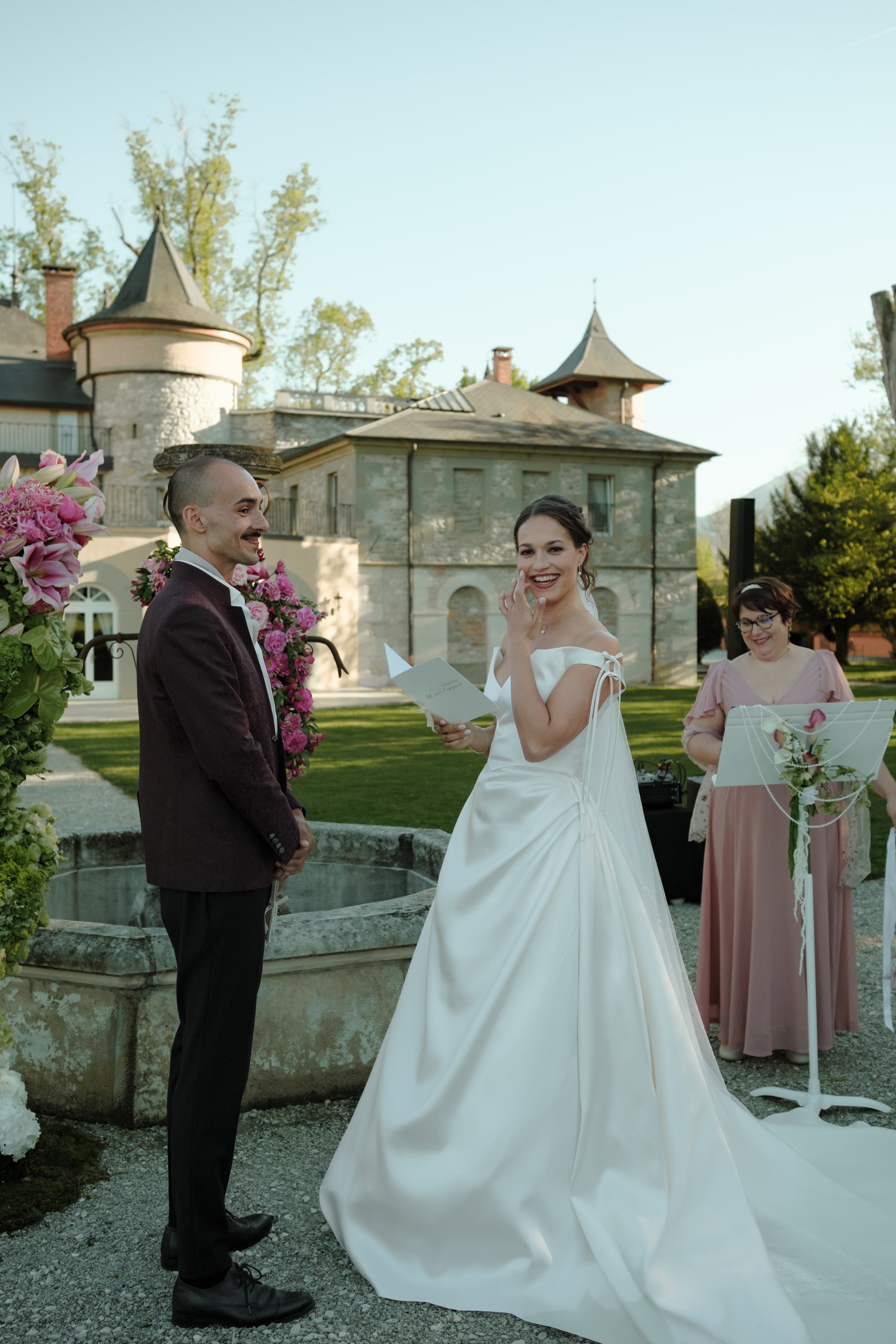 Un couple marié échange leurs vœux lors d'une cérémonie en plein air devant un château en pierre, avec une femme en robe de demoiselle d'honneur à droite.