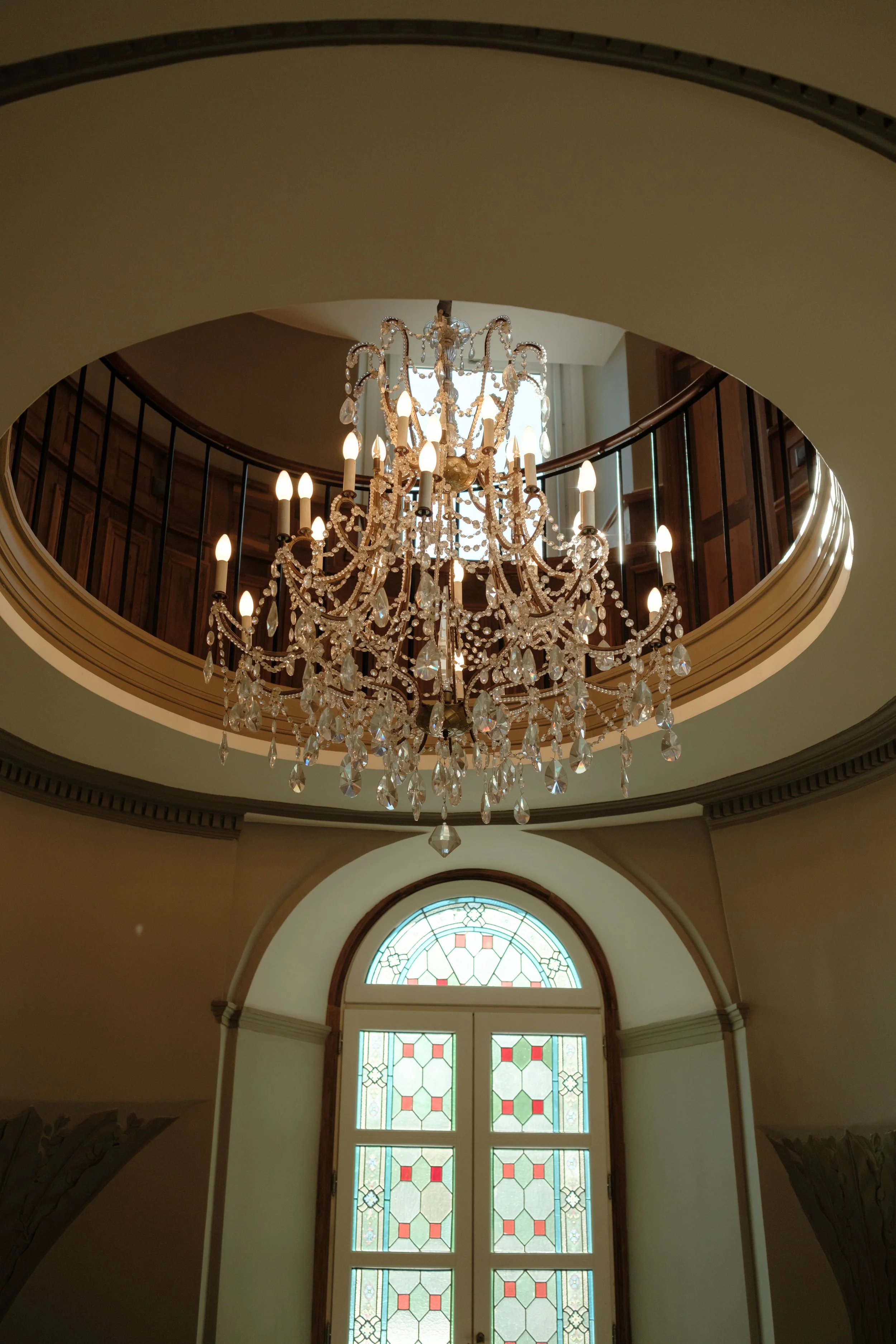 Un lustre en cristal suspendu au plafond dans un hall avec une grande fenêtre ornée de vitraux colorés.