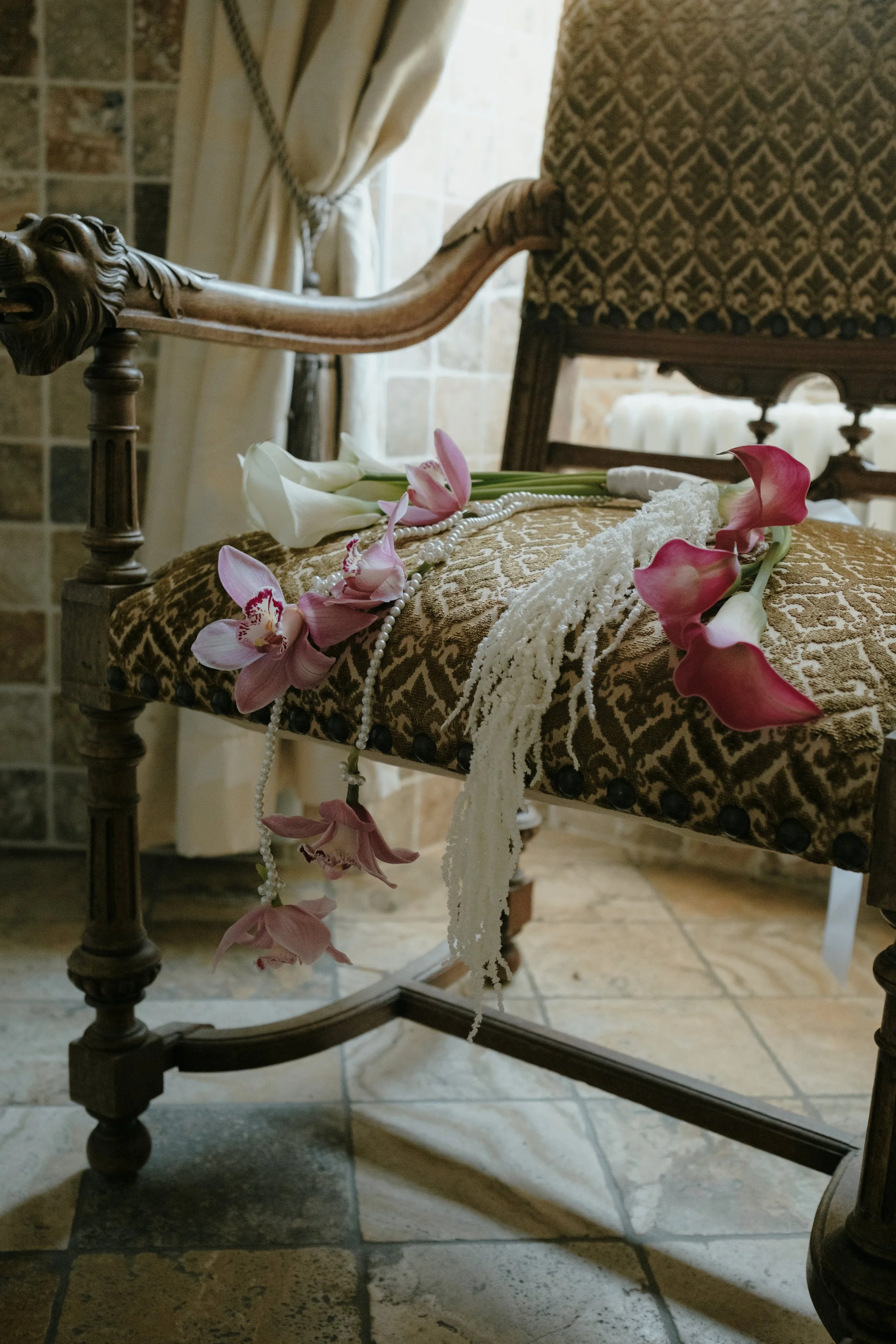 Une chaise ancienne en bois avec un tissu à motifs, des fleurs (callas roses et blanches), un collier de perles, et une serviette en dentelle sont placés dessus, dans une pièce éclairée par la lumière naturelle.