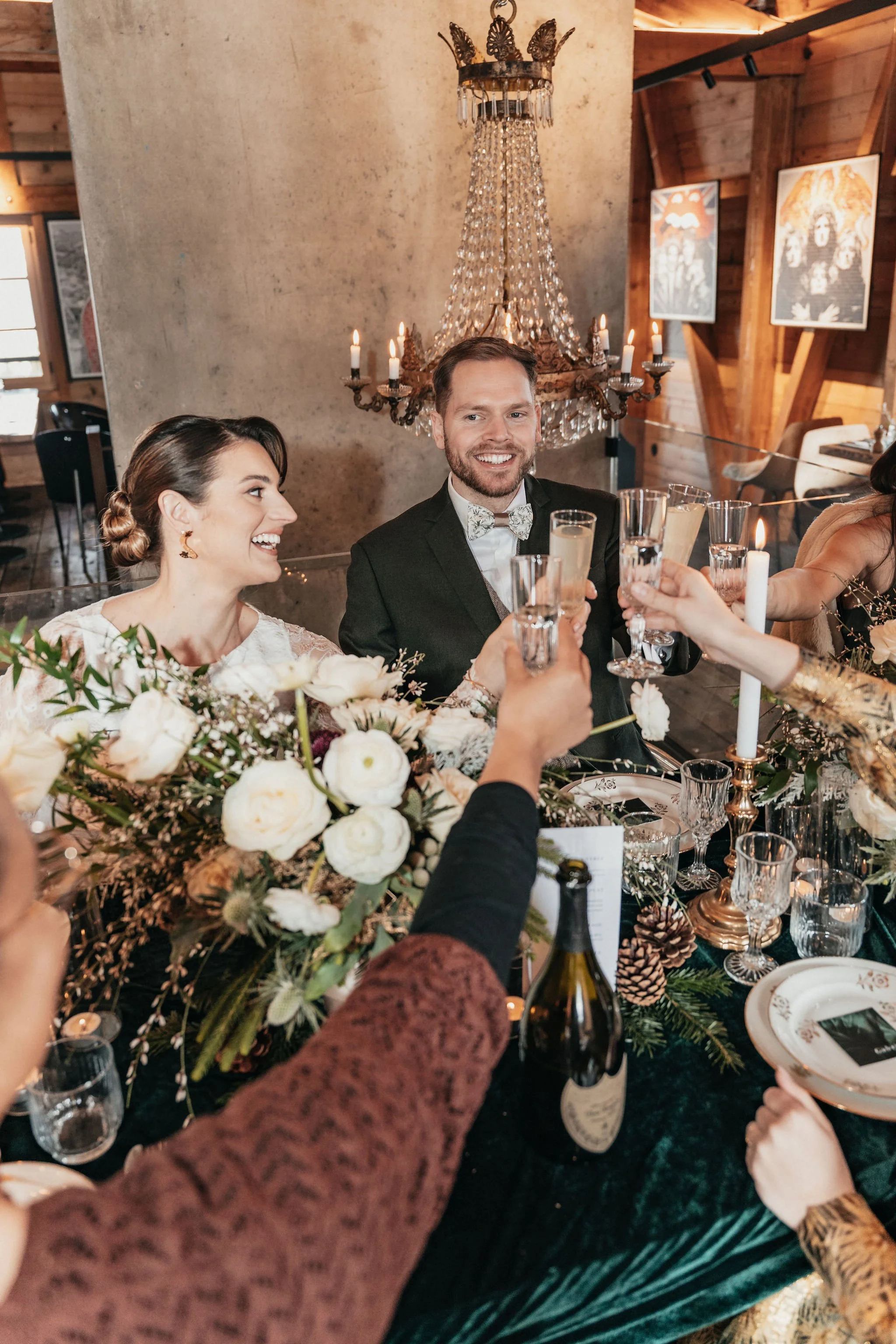 Un groupe de personnes célébrant un toast lors d'un dîner élégant, avec un couple principal au centre, autour d'une table décorée de fleurs, de bougies et de vaisselle raffinée, dans un intérieur chaleureux et chic.