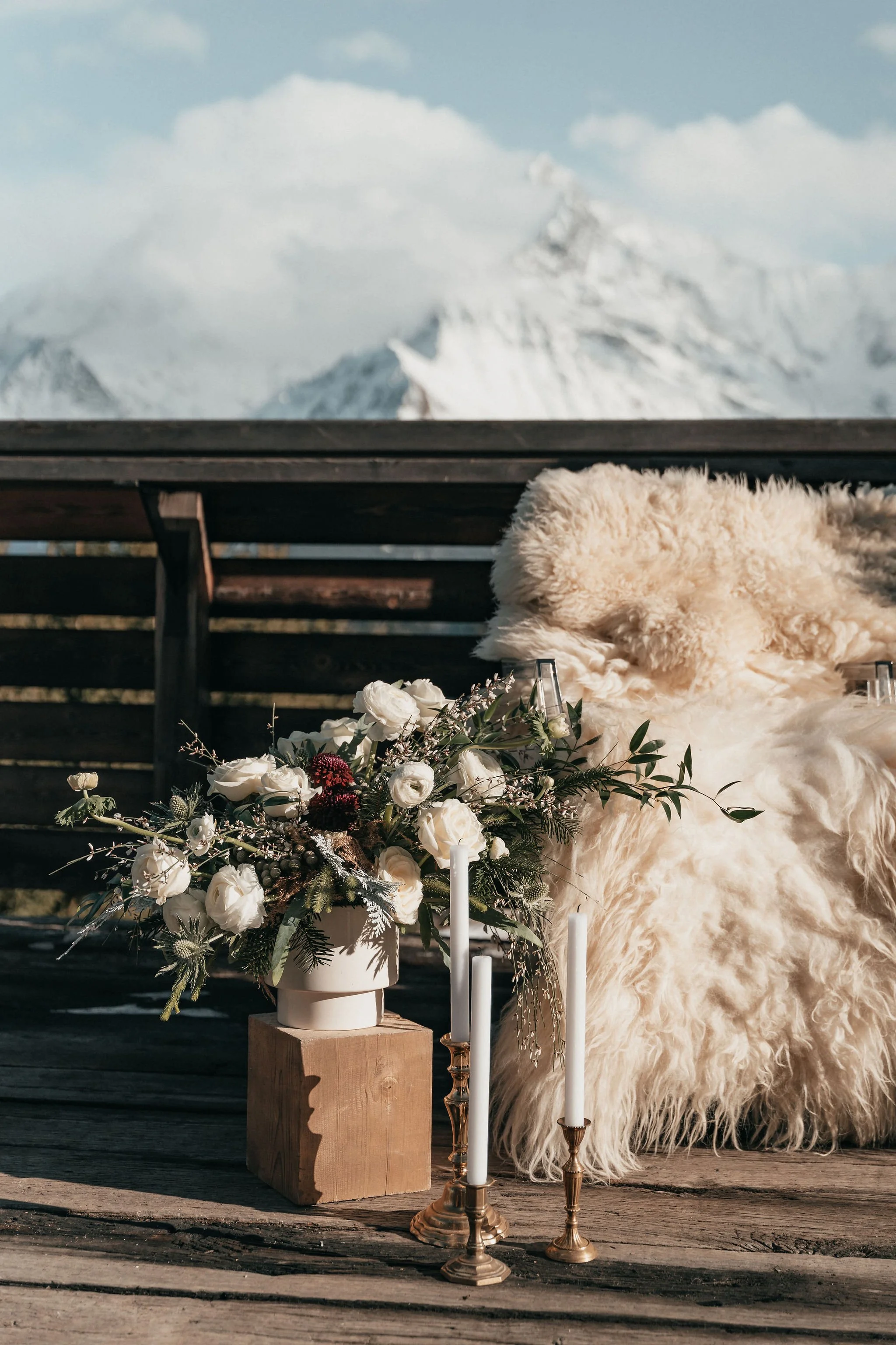 Un arrangement floral blanc avec des bougies, un tapis en laine blanche et une vue sur des montagnes enneigées en arrière-plan.