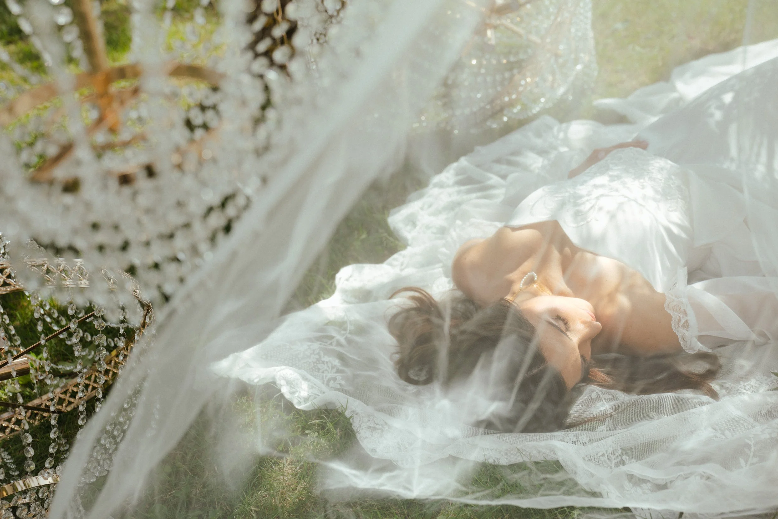 Une jeune femme en robe blanche repose dans l'herbe à l'extérieur, vue à travers une structure décorative ornée de perles ou de cristaux, créant un effet de flou artistique et romantique.
