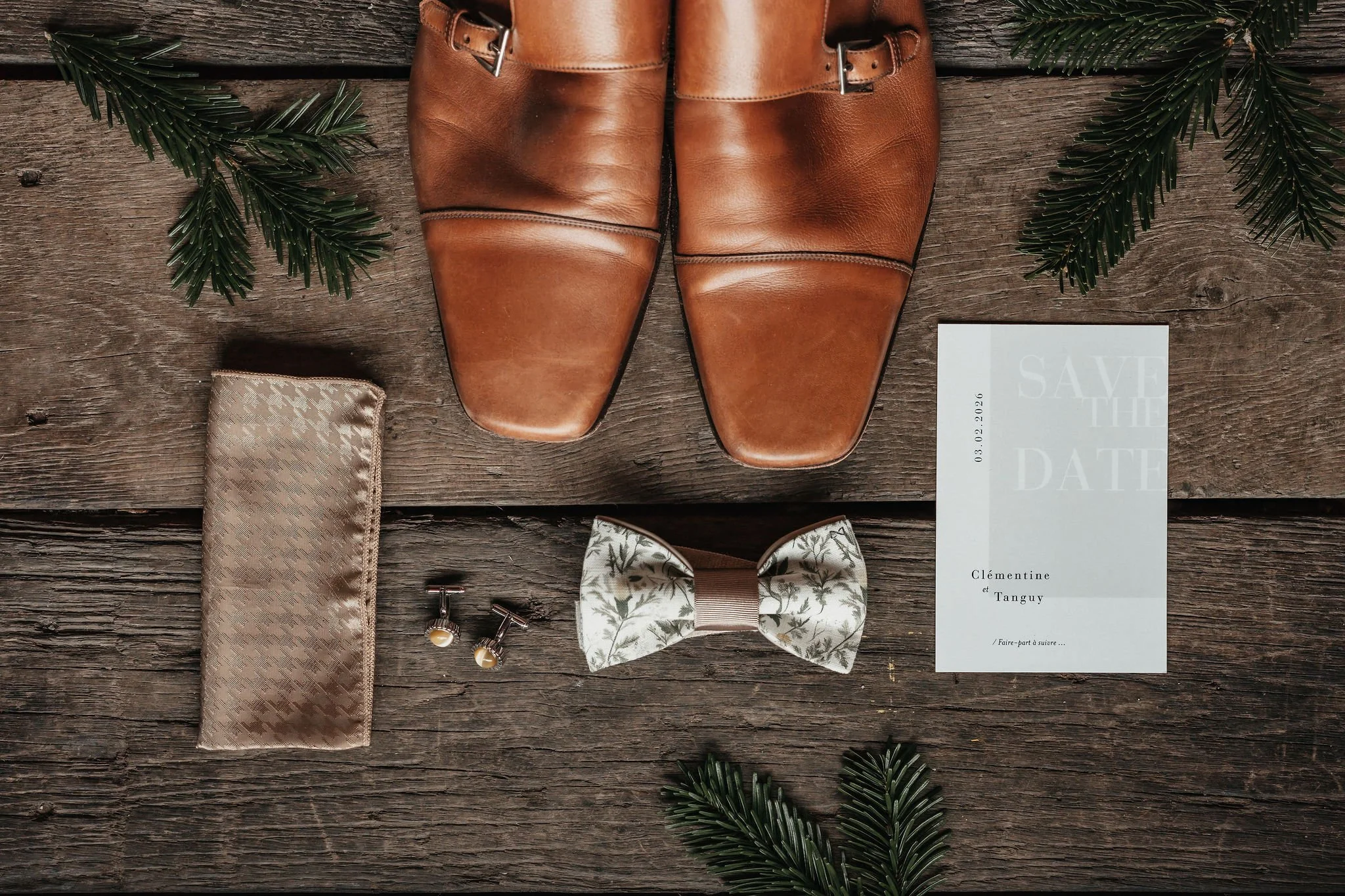 Chaussures en cuir marron, pochette beige, boutons de manchette, nœud papillon à motif floral, invitation mariage blanche, décor de sapin en branches vertes sur un fond en bois.