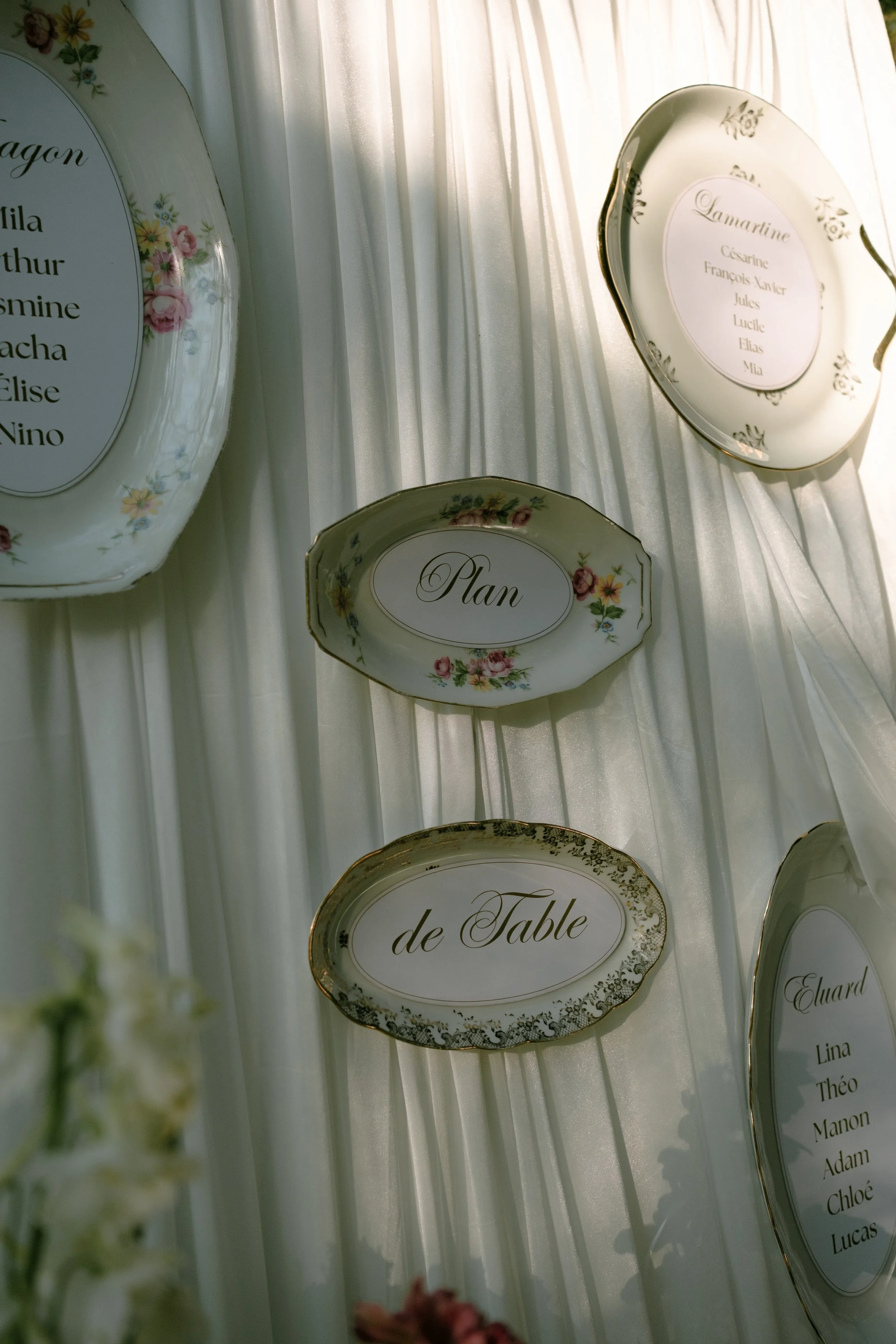 Plats en porcelaine avec des noms inscrits, probablement des places assises ou des tables pour un événement, décorée de fleurs et disposée contre un rideau blanc.