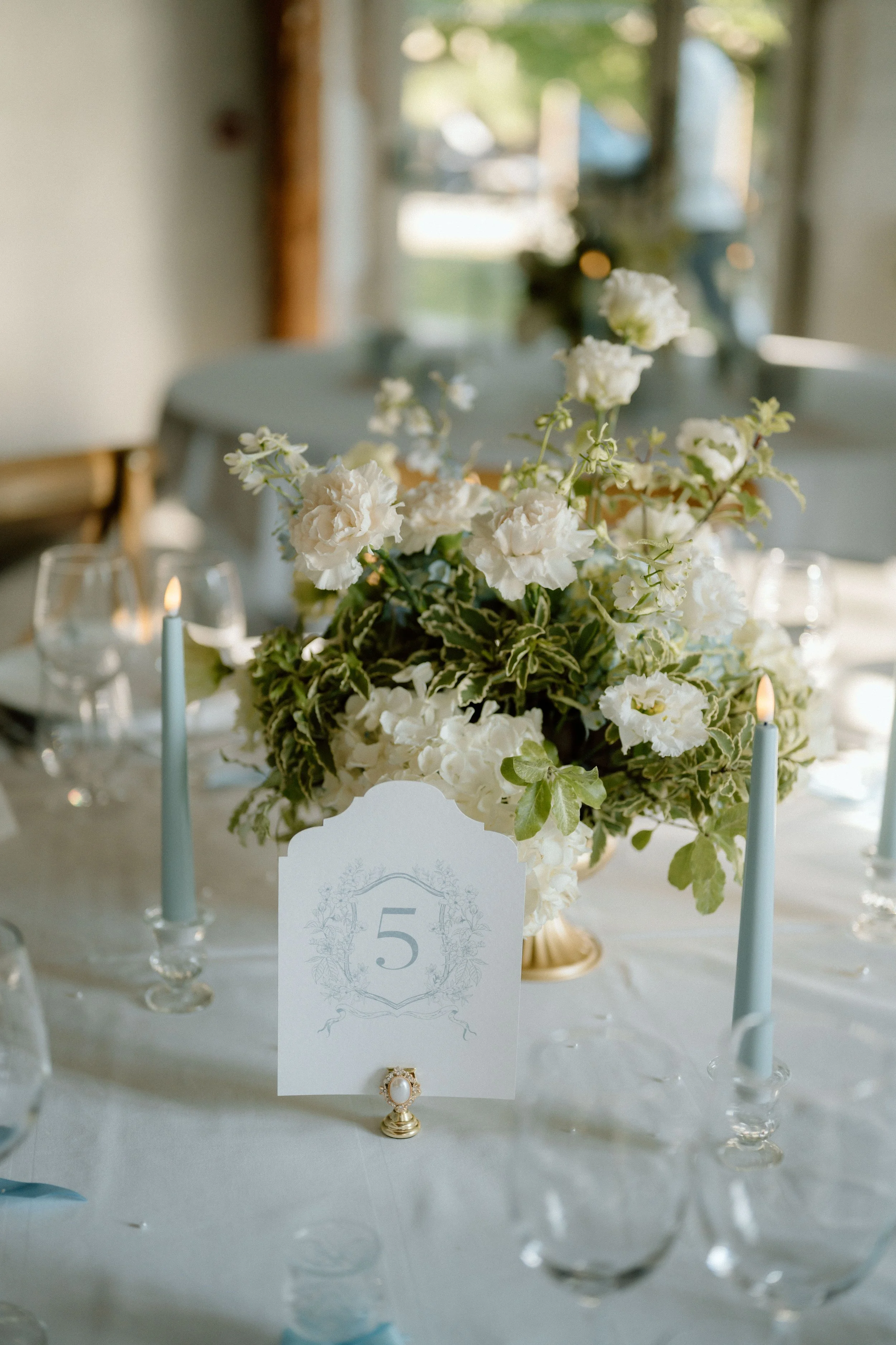Centre de table avec un bouquet de fleurs blanches, des bougies bleues, et une carte de table portant le numéro 5, pour un événement ou une réception dans une pièce lumineuse.
