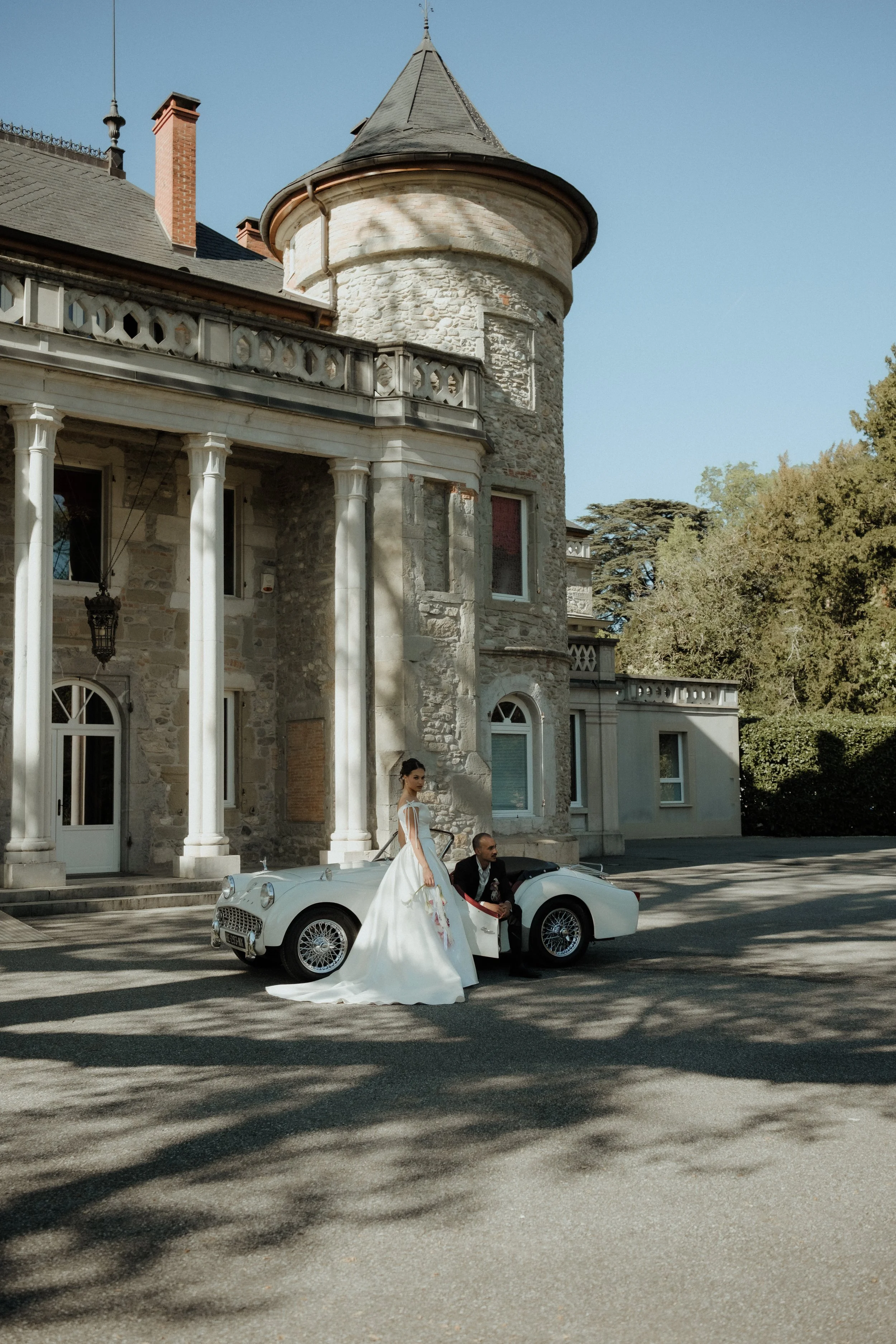 Une femme en robe de mariée blanche et un homme en costume noir posent à côté d'une voiture de sport blanche devant un bâtiment en pierre avec une tour conique.