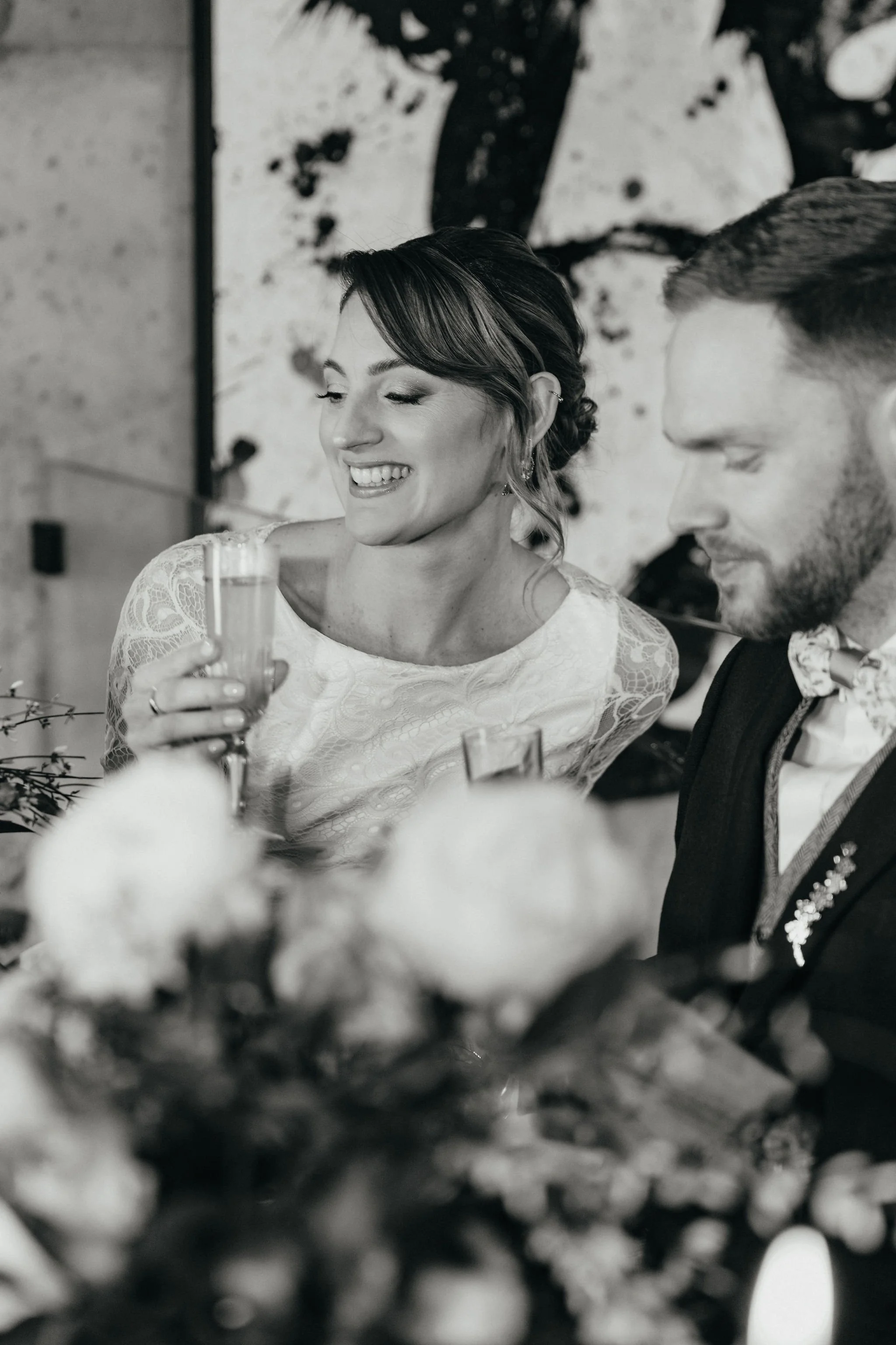 Une femme souriante avec une robe en dentelle tient un verre lors d'une célébration, avec un homme à côté, dans un décor artistique.