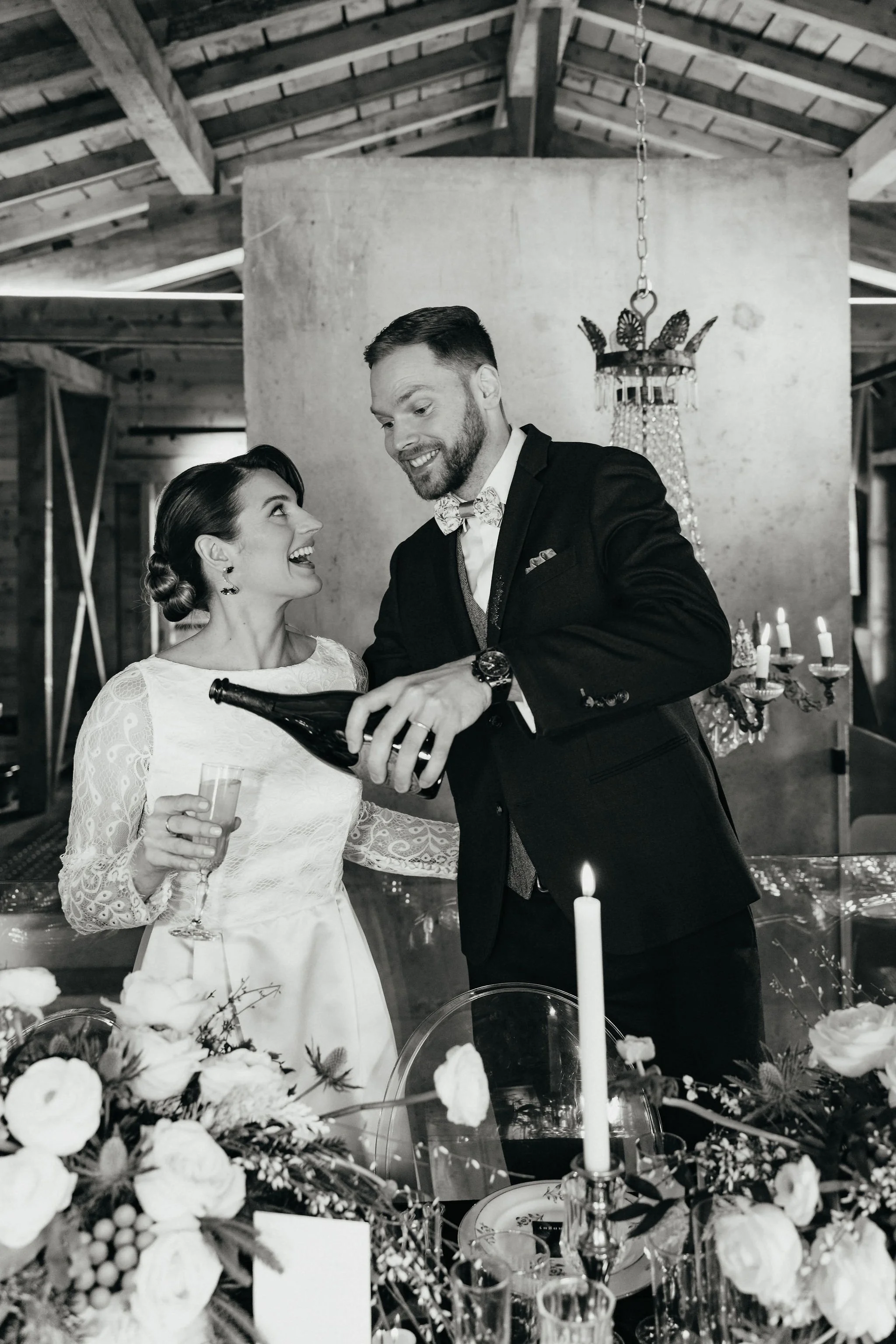 Un couple marié partage un toast lors de leur mariage, en pleine coupe de champagne, dans une salle décorée avec des fleurs et une chandelier.
