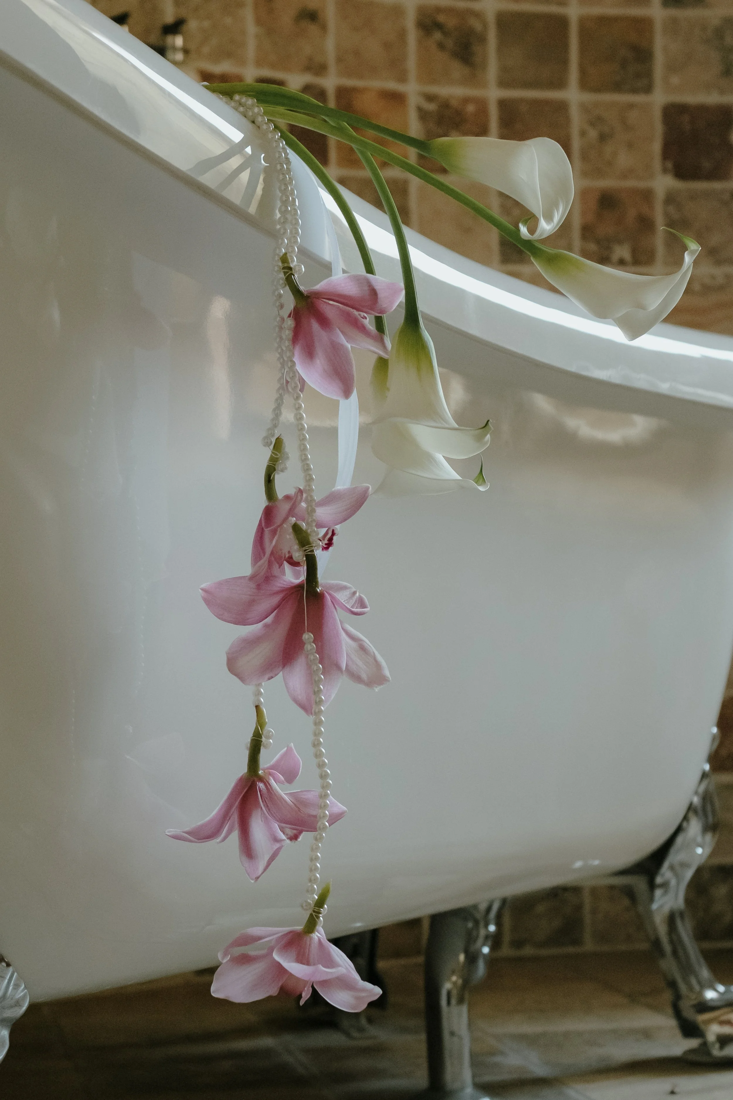 Des fleurs blanches et roses pendantes, décoratives, suspendues à une perle blanche le long d'une baignoire en porcelaine blanche, avec un mur en briques en arrière-plan.