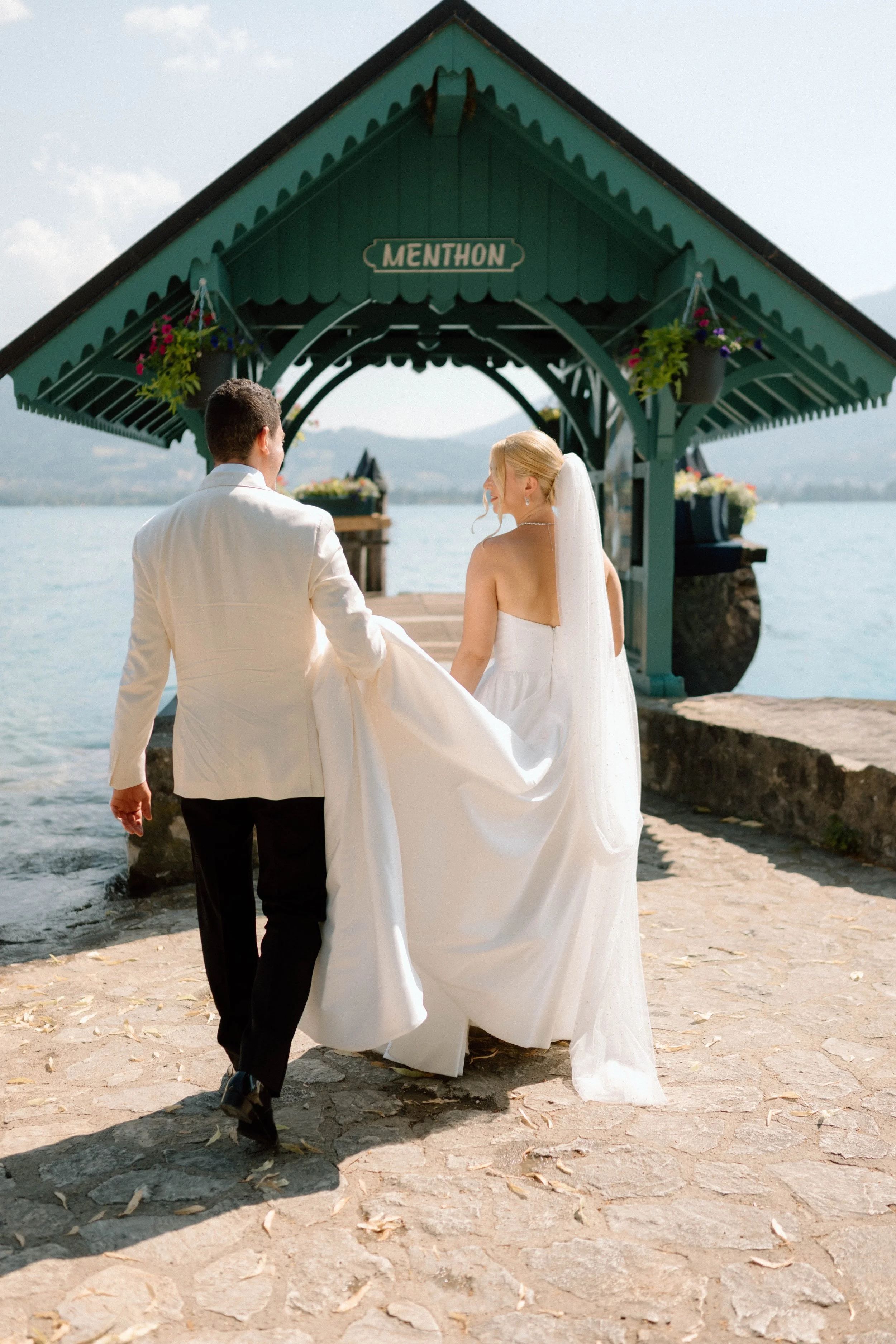 Un couple en mariage marchant sur une jetée au bord de l'eau, avec un pavillon en bois marqué "Menthon" et des montagnes en arrière-plan.