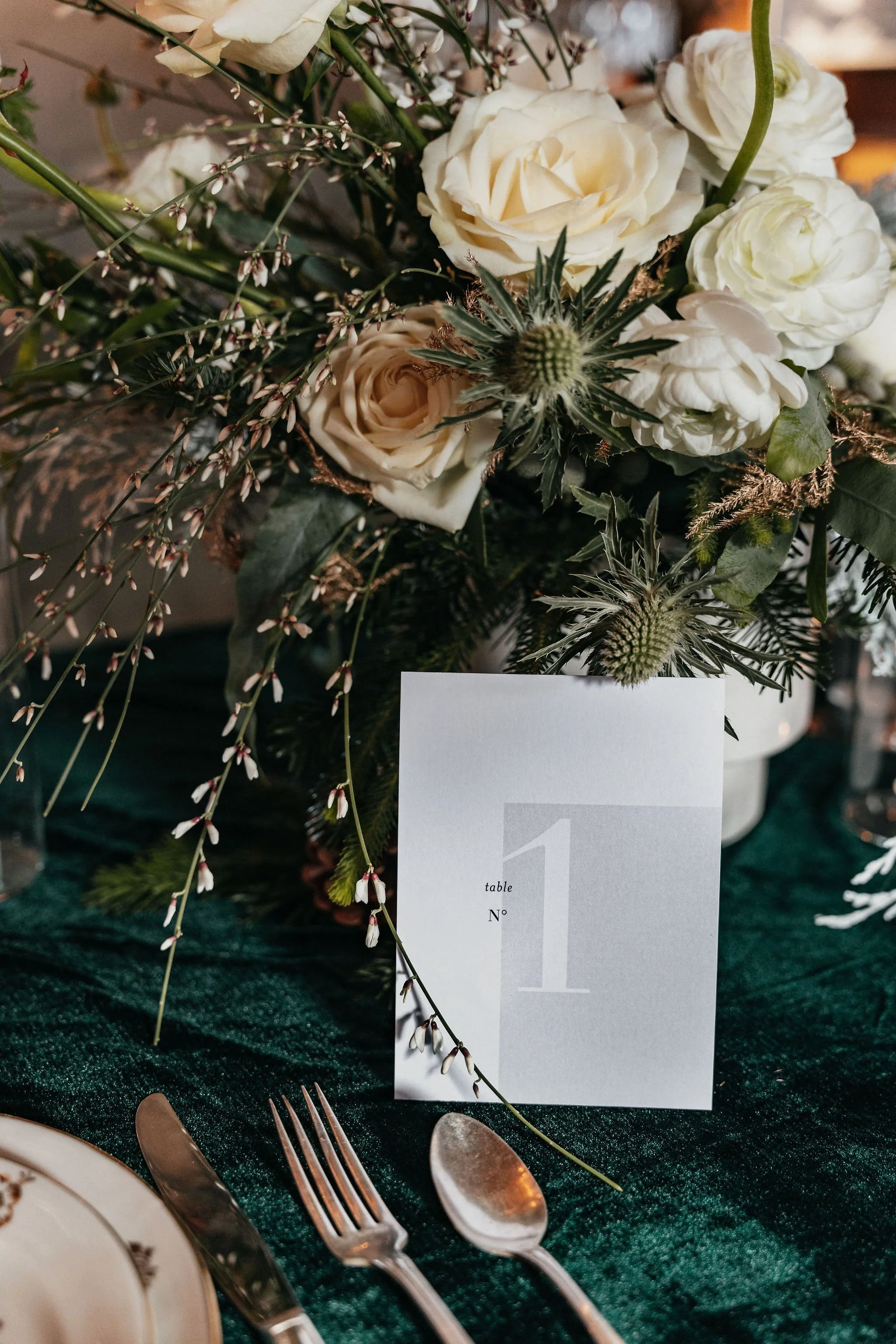Un centre de table floral avec des roses blanches, des giroflées, des feuilles vertes et des éléments de décoration, sur une nappe verte foncée avec une carte de table indiquant le numéro 1.
