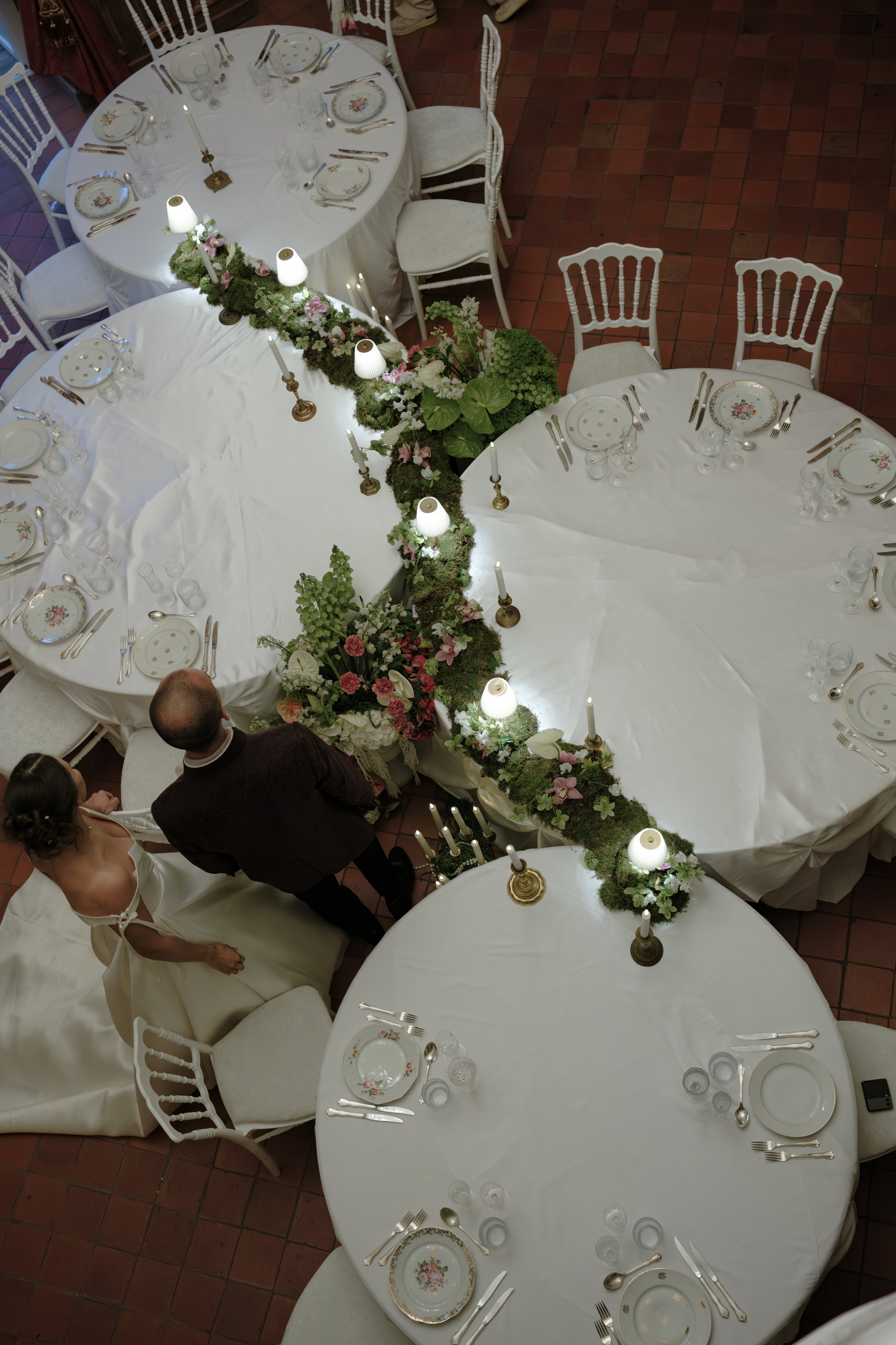 Table dressée pour un événement ou un repas, décorée avec des fleurs, des bougies, des assiettes en porcelaine avec motifs floraux, et de la vaisselle en verre. La table est entourée de chaises blanches.
