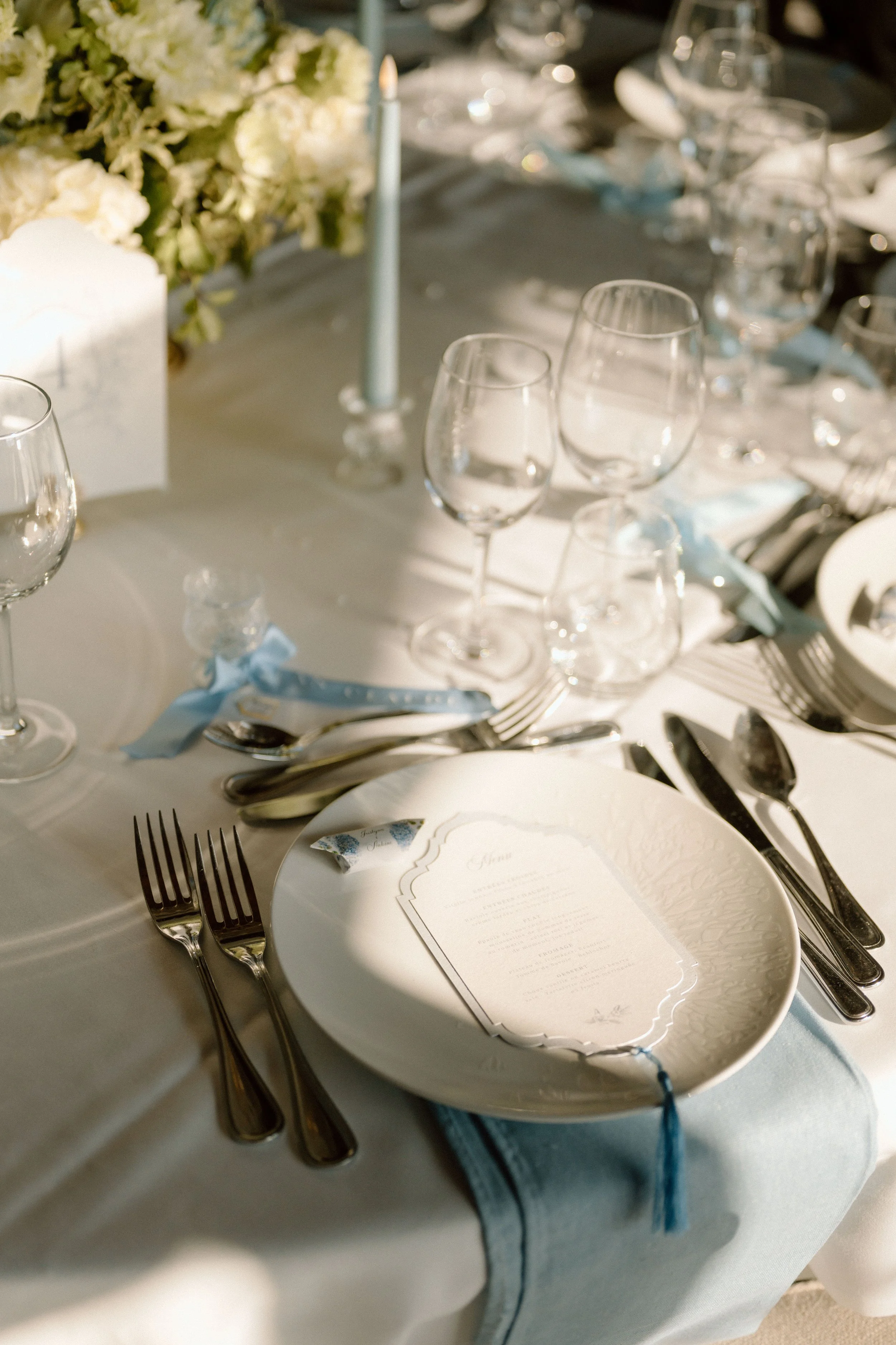 Table de mariage élégante avec des verres à vin, des couteaux, des fourchettes, une assiette avec une carte de menu, des bougies et une décoration florale blanche.