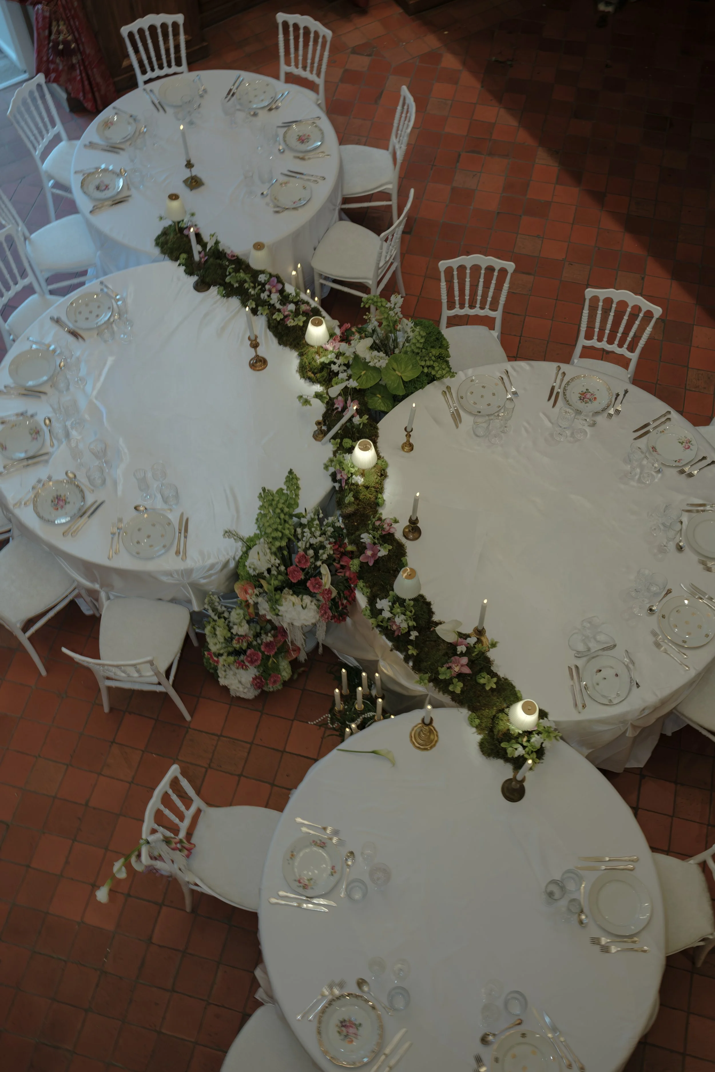 Tables de banquet décorées avec de la vaisselle fine, des bougies et des arrangements floraux dans une salle avec un sol en carreaux rouges.