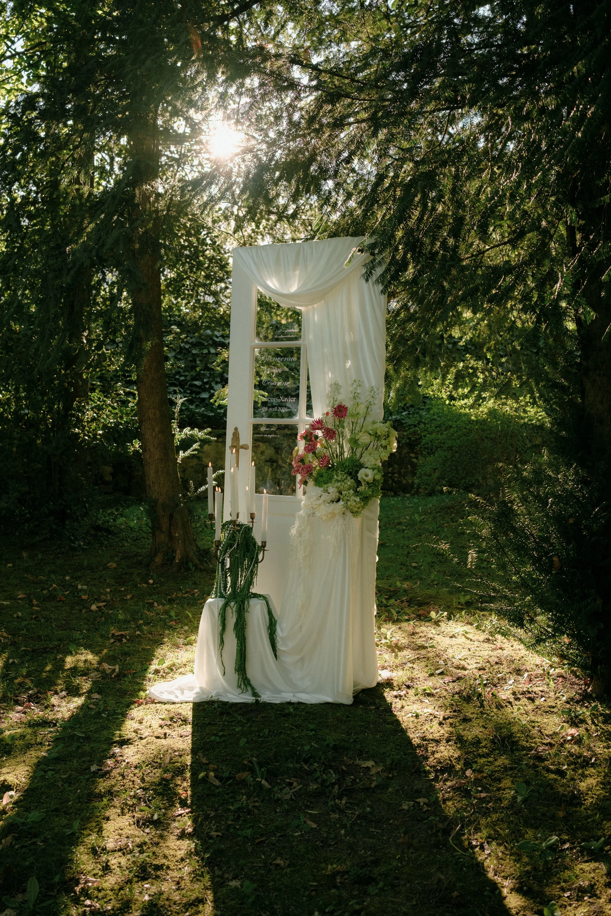 Décoration de mariage dans un jardin avec un cadre blanc et des fleurs, baigné de lumière du soleil entre les arbres.