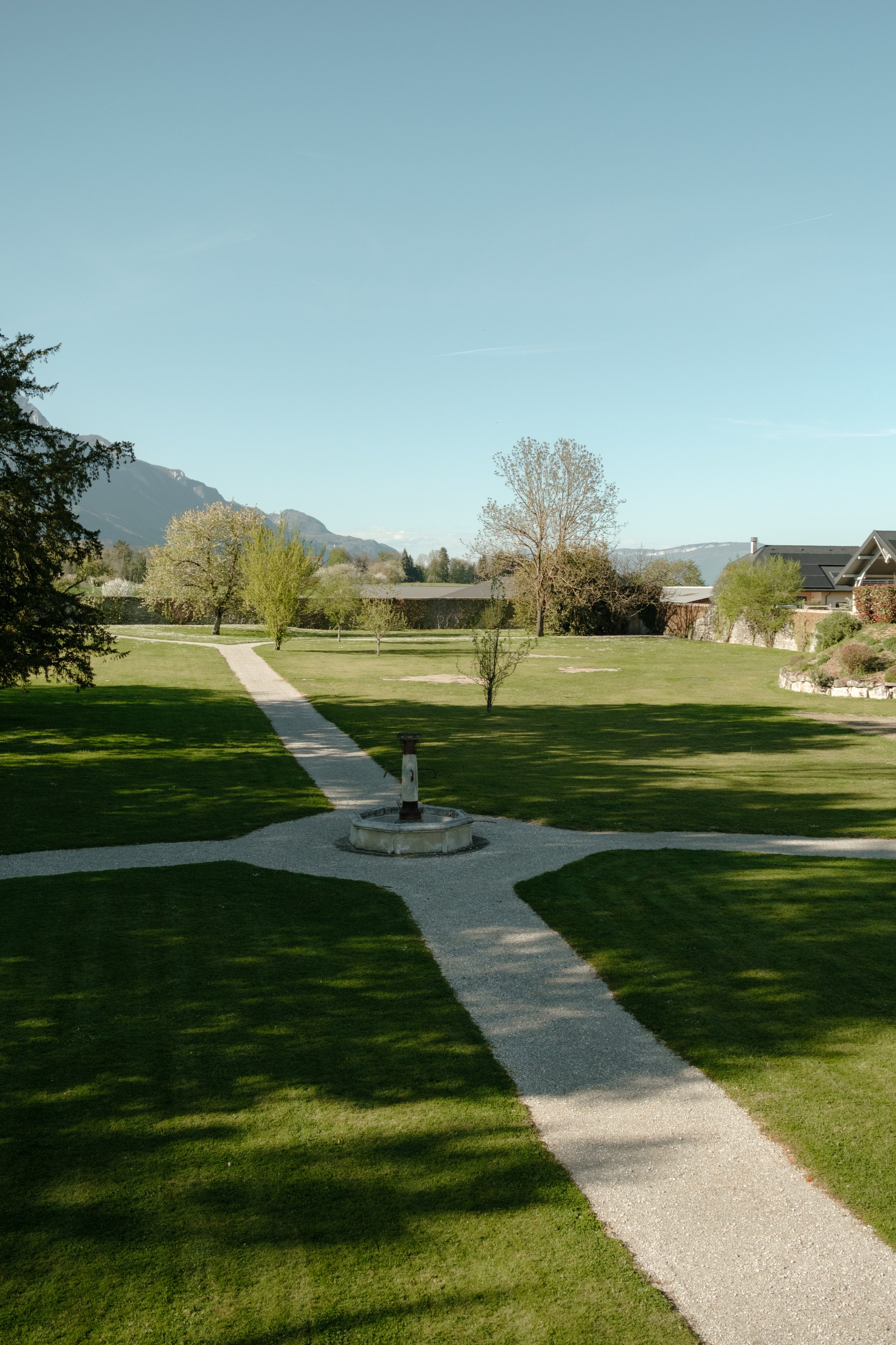 Un jardin avec des arbres, une fontaine, un chemin en gravier, et une maison en arrière-plan.