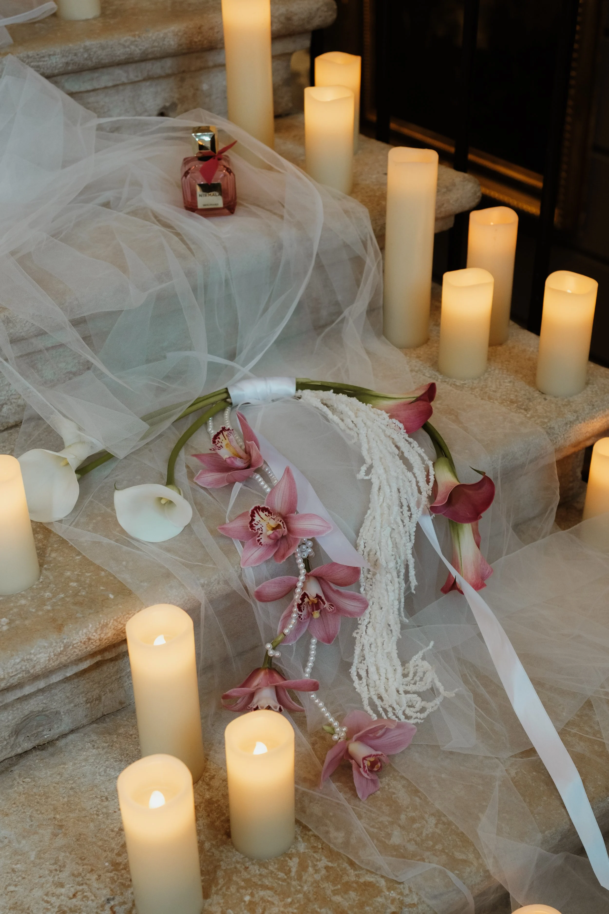 Arrangements florales avec des fleurs d'orchidée roses et blanches, tulle blanc, et plusieurs bougies sur un rebord en pierre, probablement pour une cérémonie ou un mariage.