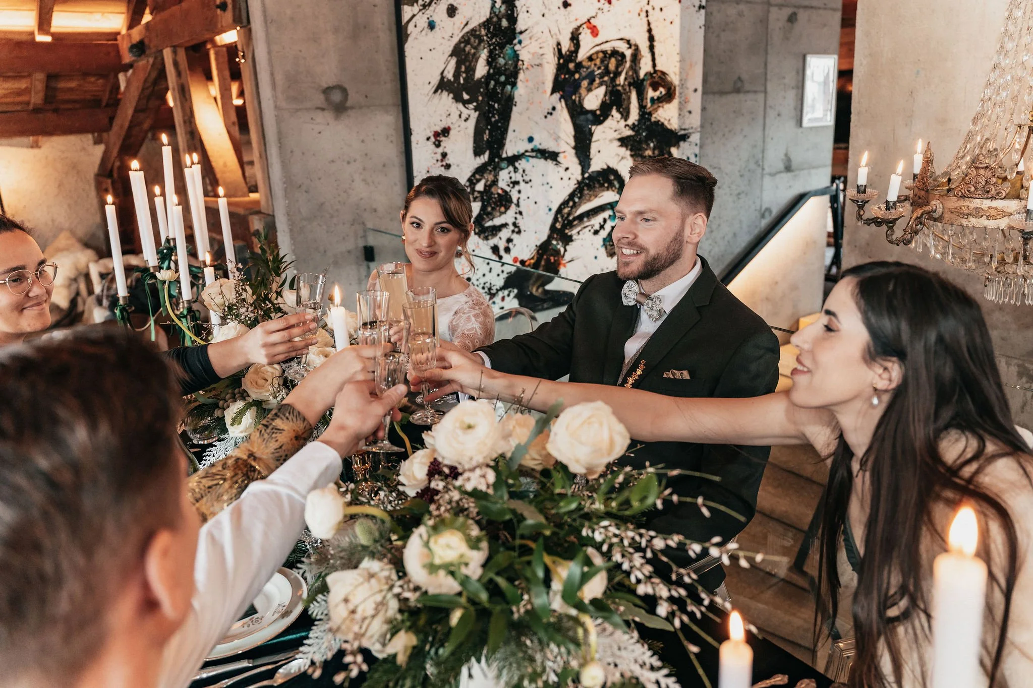 Groupe de personnes lors d'une célébration, levant des verres pour un toast, autour d'une table décorée avec des fleurs et des bougies, dans un espace intérieur élégant.