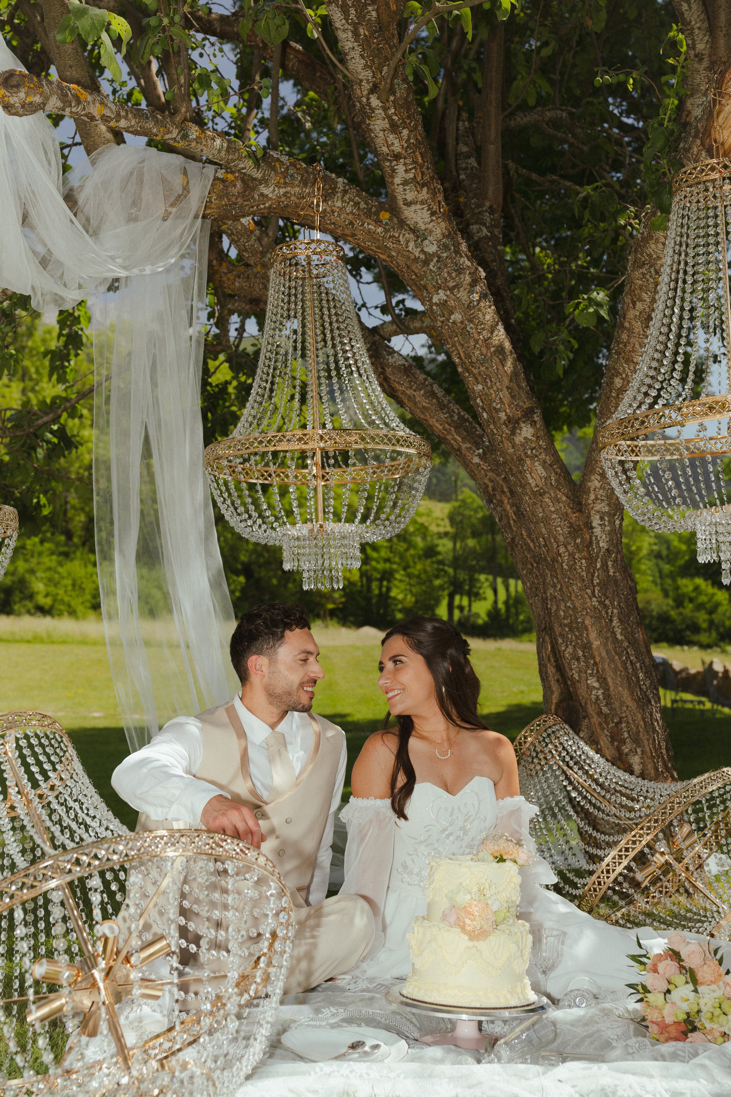 Un couple marié assis à une table de mariage en plein air, souriant et regardant l'un l'autre, avec un gâteau de mariage blanc avec des fleurs pastel, des chandeliers et une décoration élégante en cristal et en or, sous un arbre avec des chandeliers 
